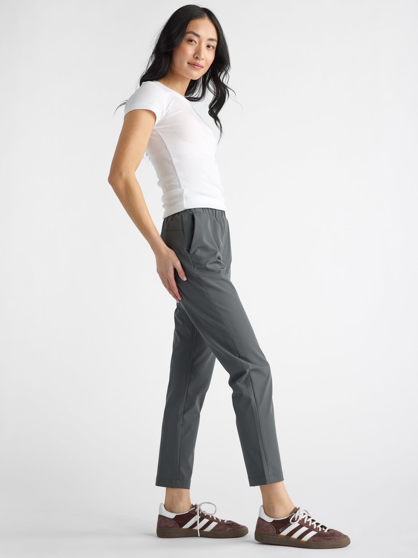 A woman stands against a plain background, wearing a white t-shirt, Women's Always Cropped Pant by Cozy Earth in gray, and maroon and white athletic shoes. They pose with one hand in their pocket, slightly turned to the side. |Color:Coal