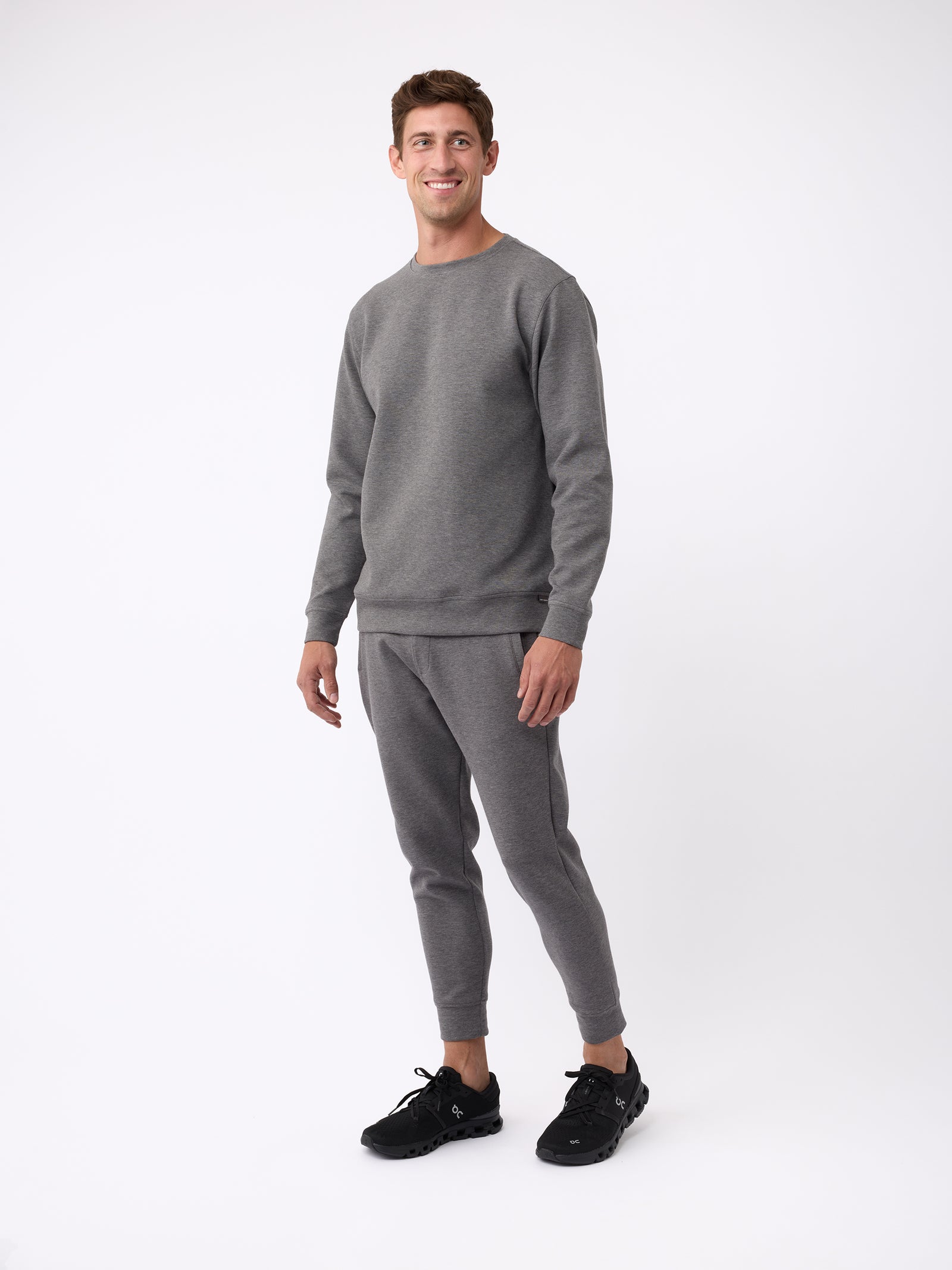 A man is smiling and wearing a Cozy Earth matching gray Men's StretchTech Crewneck sweatsuit with black sneakers. He is standing against a plain white background. 