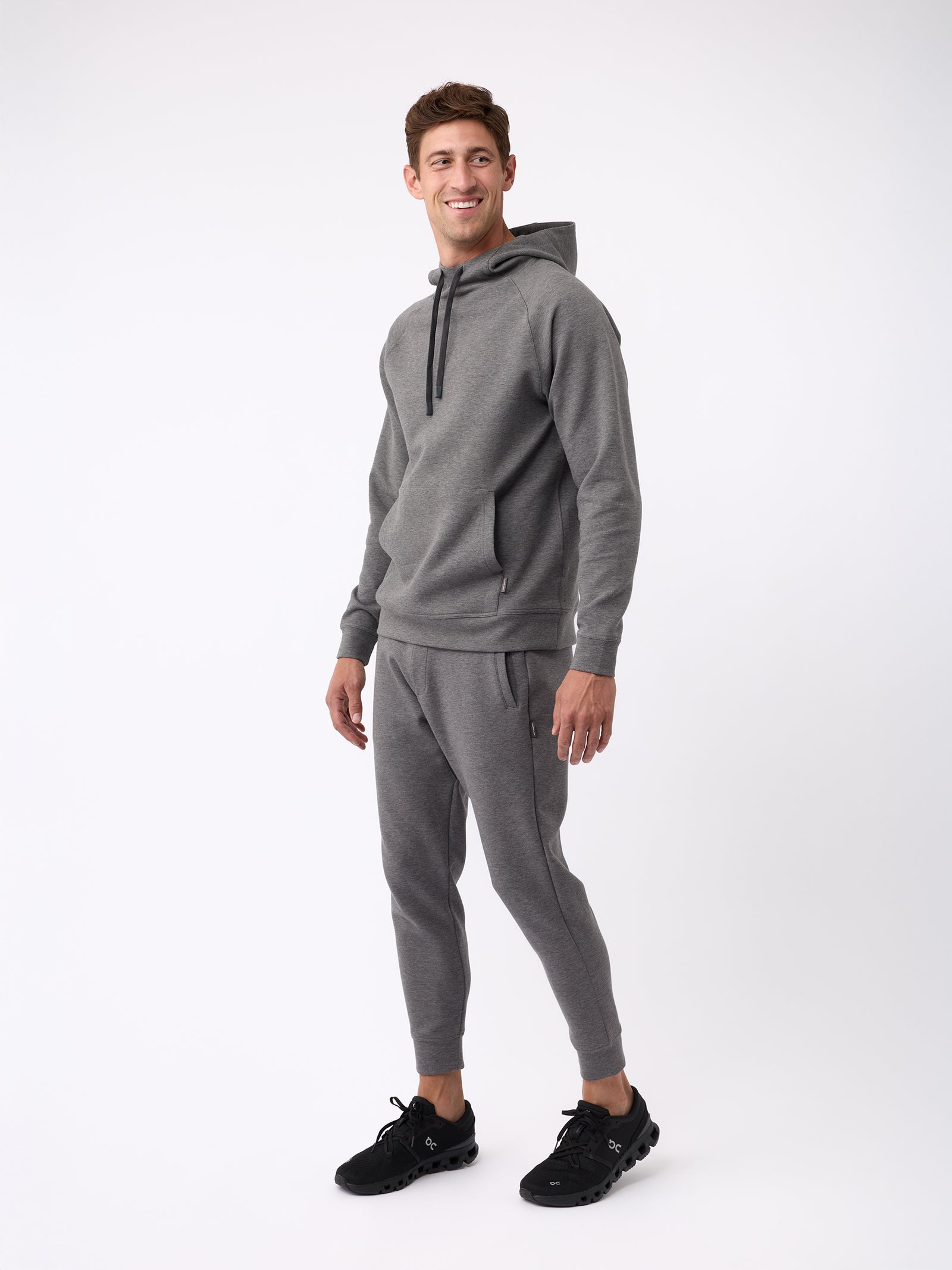 A person is smiling while wearing a Cozy Earth Men's StretchTech Hoodie in gray along with matching joggers, standing against a plain white background. They are also wearing black sneakers. 