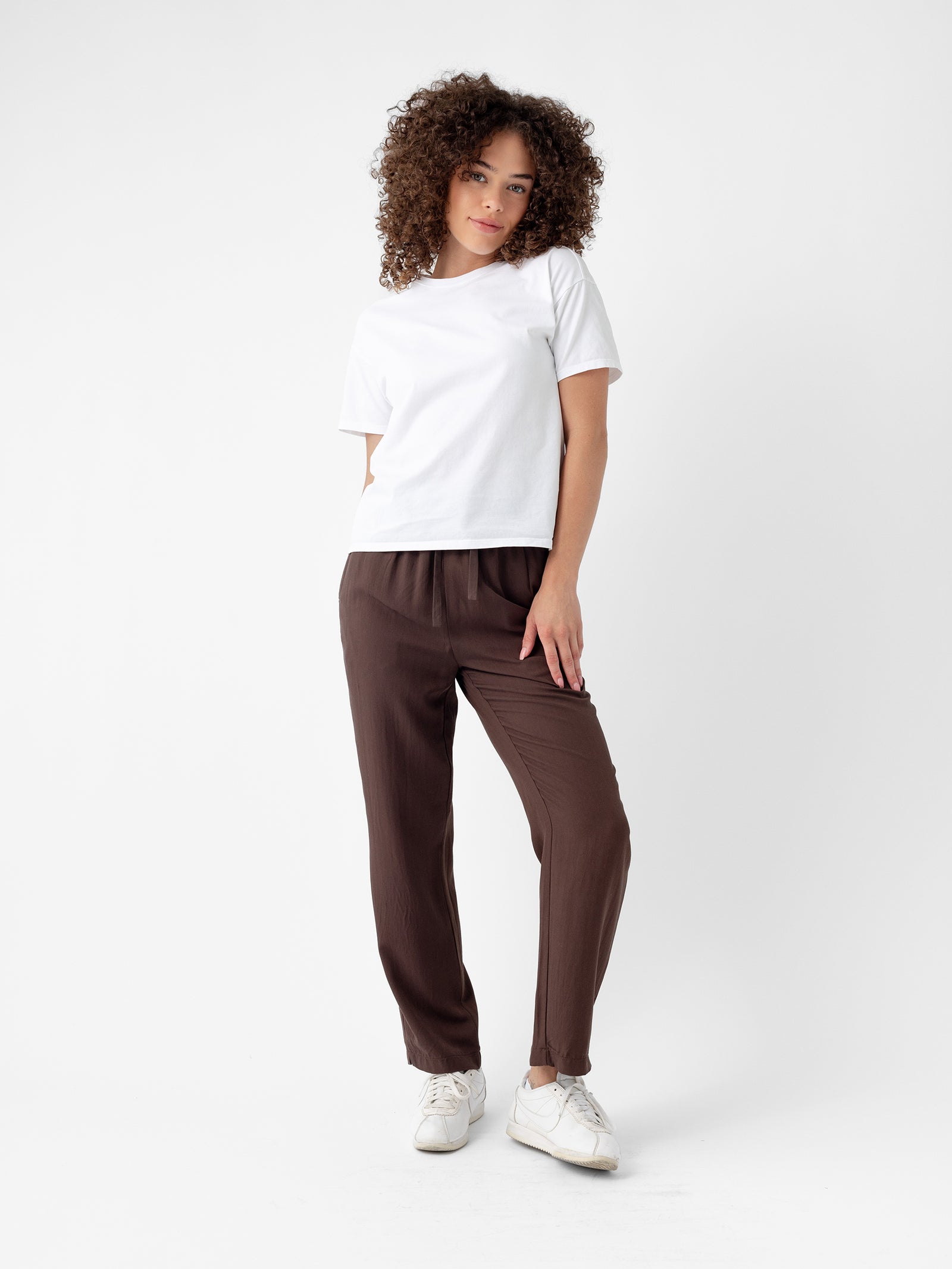 A person with curly hair stands against a plain white background, wearing a white T-shirt, Women's Sunset Cropped Pant from Cozy Earth, and white sneakers. They have a relaxed posture with one hand in their pocket and a calm expression. 