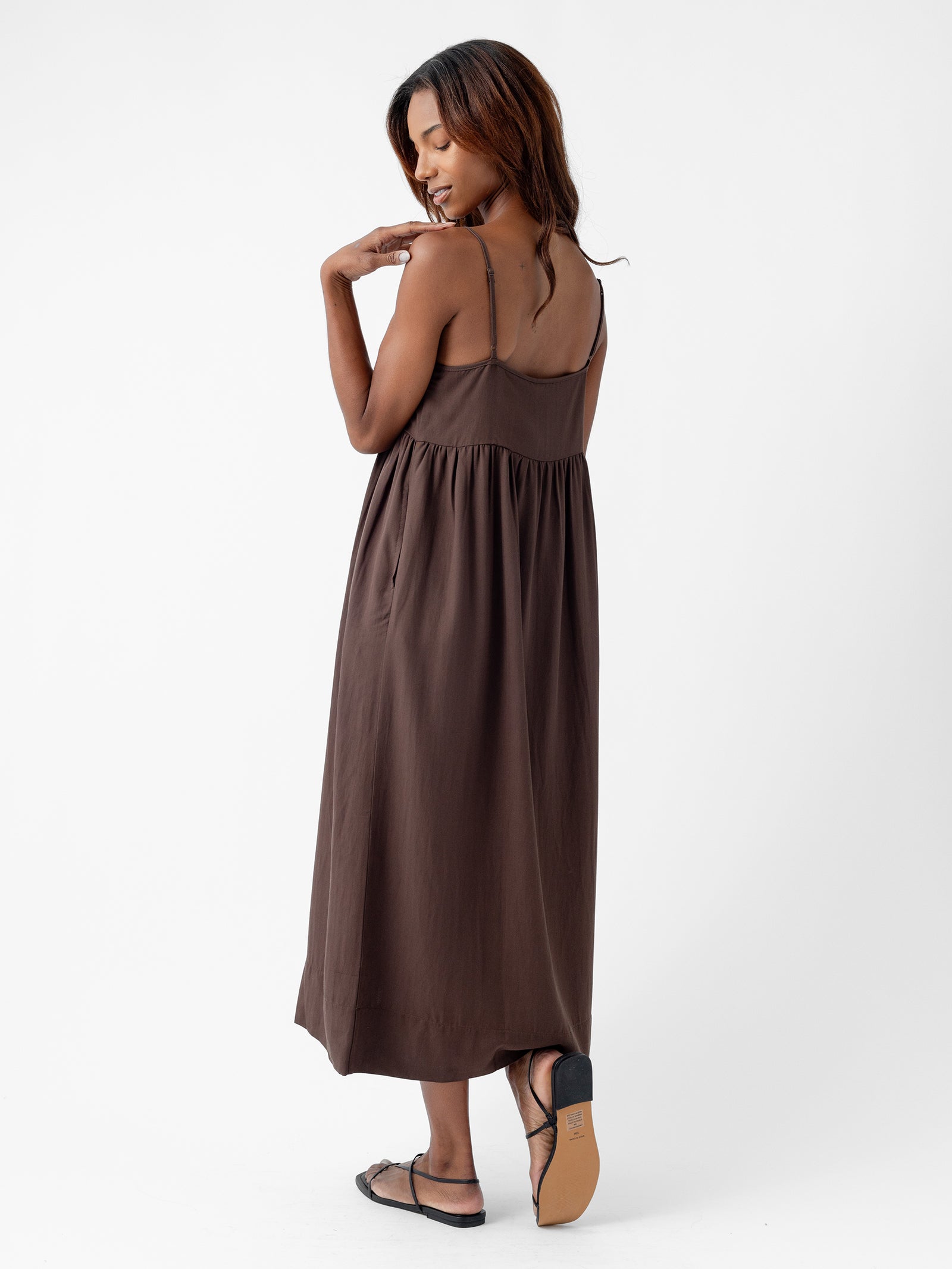 A woman with long hair touches her shoulder as she looks down, wearing Cozy Earth's Women's Sunset Gathered Dress, a sleeveless, brown maxi dress with spaghetti straps and black sandals. She is standing on a white background, posing with one foot slightly raised. 