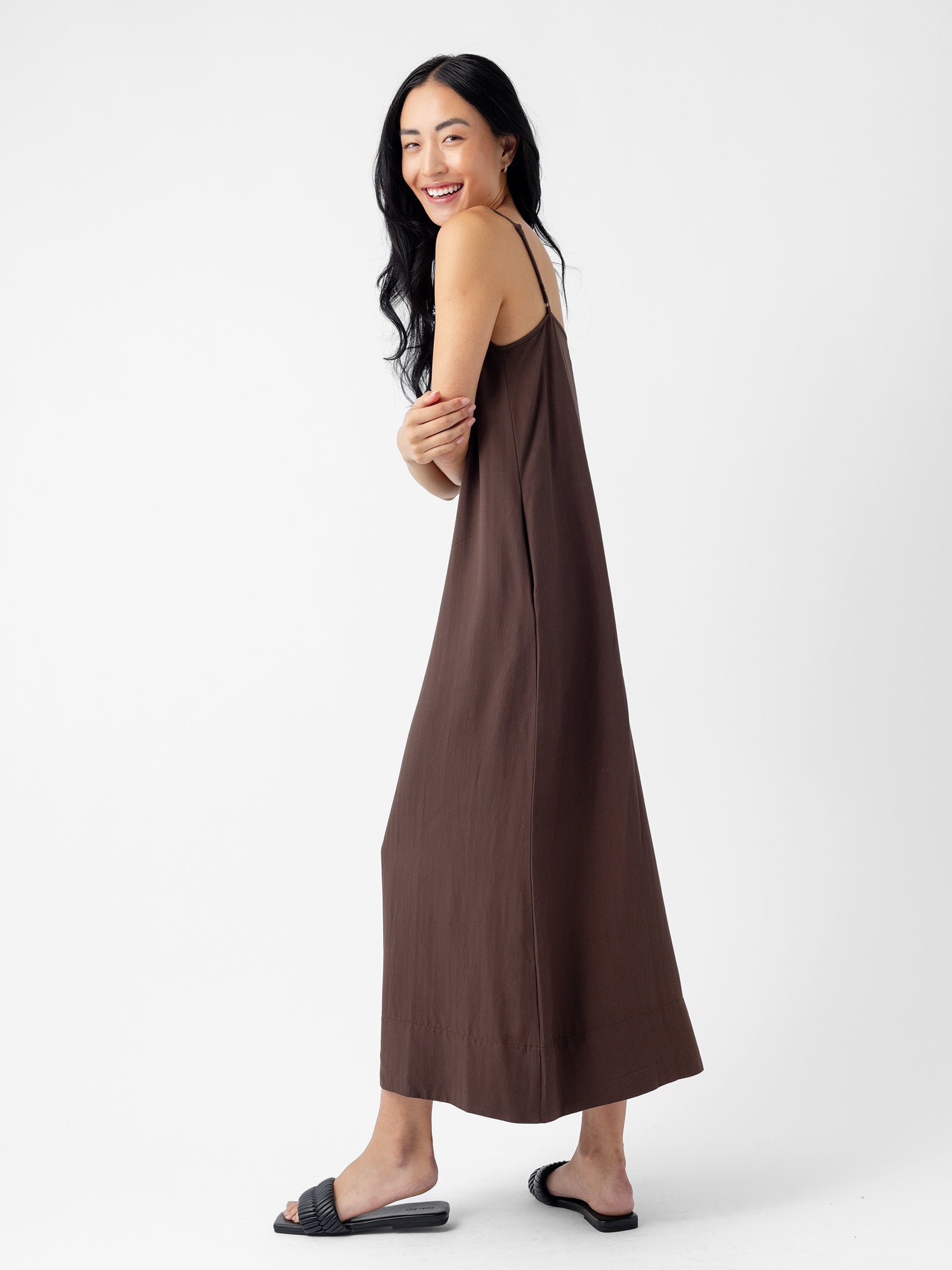 A person with long black hair smiles while wearing the Women's Sunset Slip Dress by Cozy Earth in a loose-fitting brown, sleeveless design and black slide sandals. They are standing sideways, looking over their shoulder against a plain white background. 