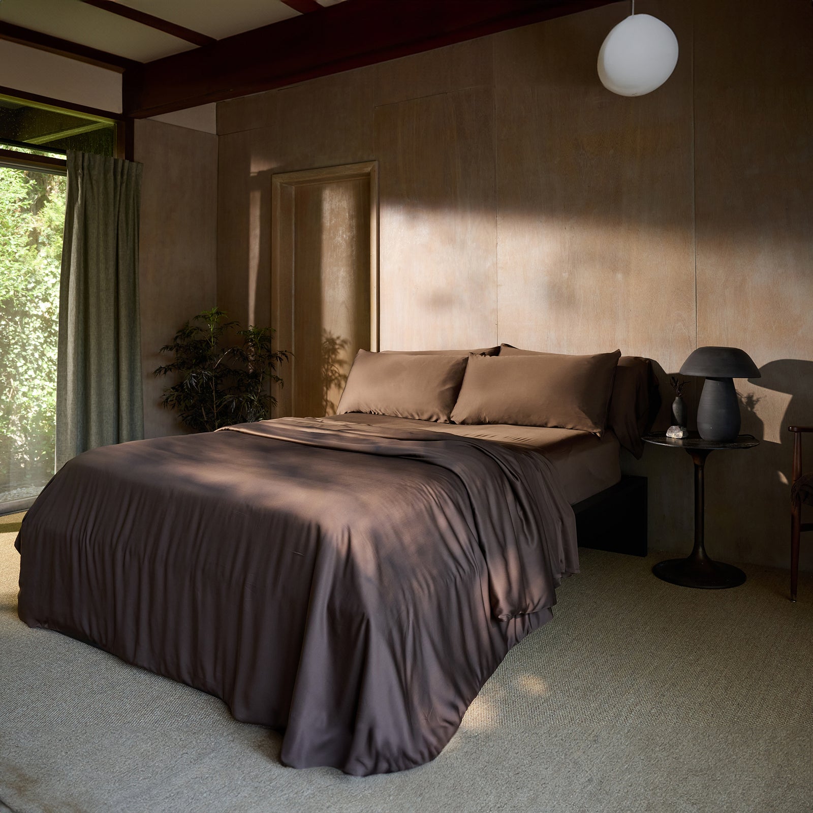 A serene bedroom with a large bed adorned with Cozy Earth's Bamboo Duvet Cover in a smooth, chocolate-brown color. The room features a neutral color palette, a nightstand with a dark lamp, a lush potted plant in the corner, curtains, and soft natural light filtering in from the window. 