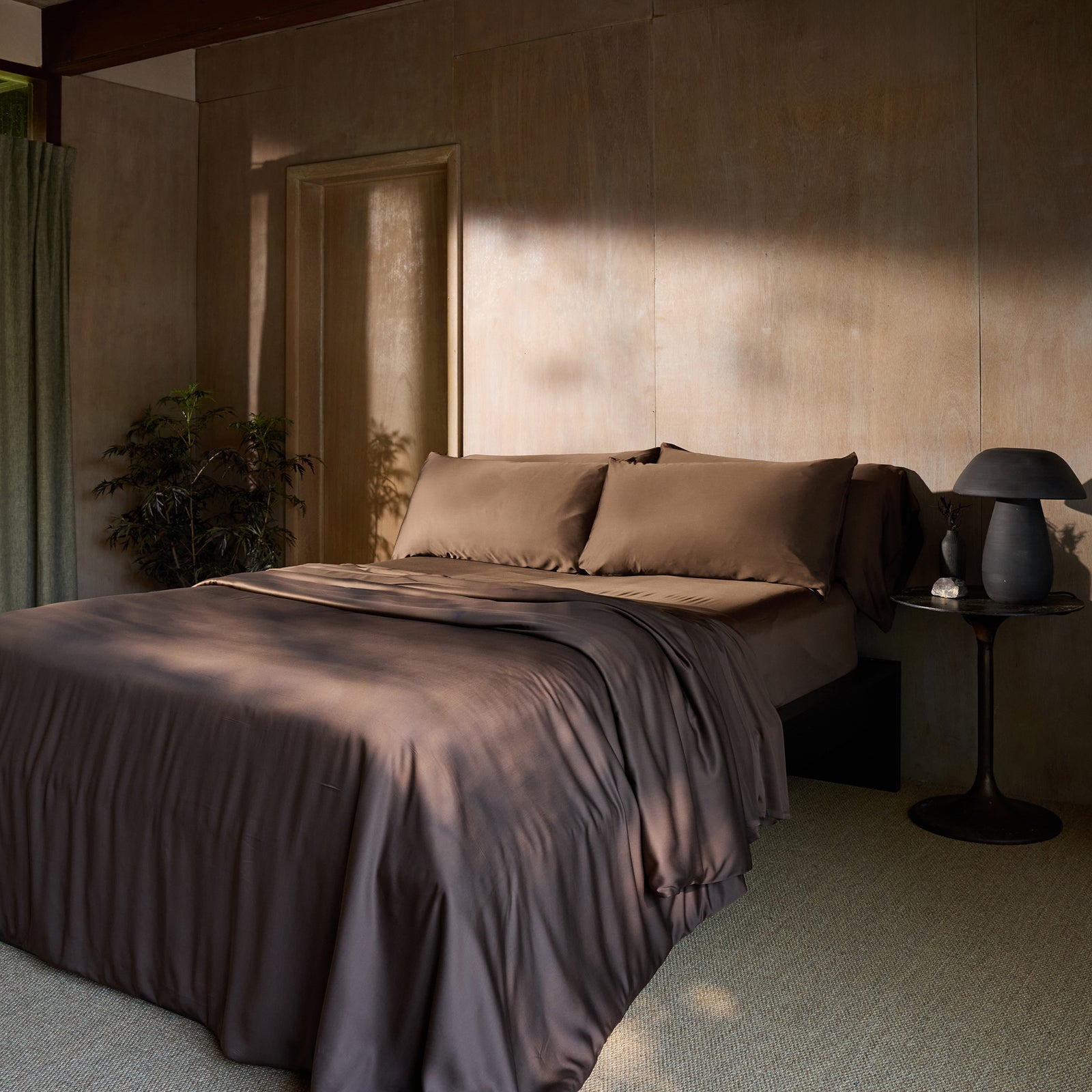 A sunlit bedroom featuring a neatly made bed with Cozy Earth's Bamboo Fitted sheet in brown and matching pillows. There's a small, round bedside table on the right with a dark lamp and a decorative object. A green plant stands in the corner, and the room has wooden walls and a beige carpeted floor. 