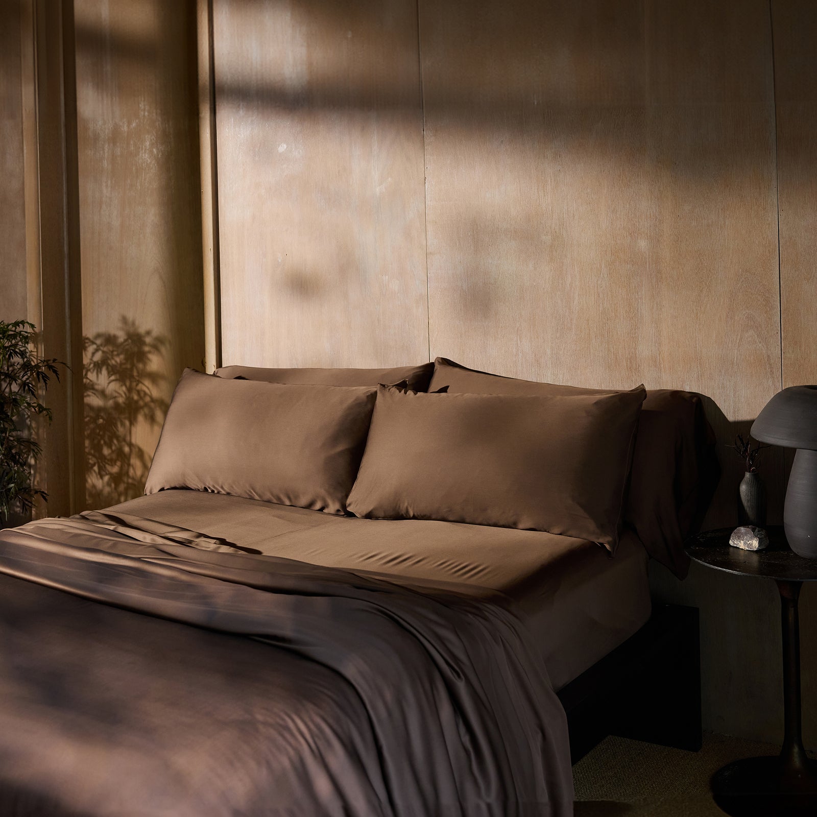 A neatly made bed with Cozy Earth's Bamboo Flat Sheet and matching pillowcases in brown is situated against a wooden wall. Soft, natural light casts gentle shadows on the bed and wall. A small round table next to the bed holds a dark, modern lamp and a decorative item. A potted plant is partially visible. 