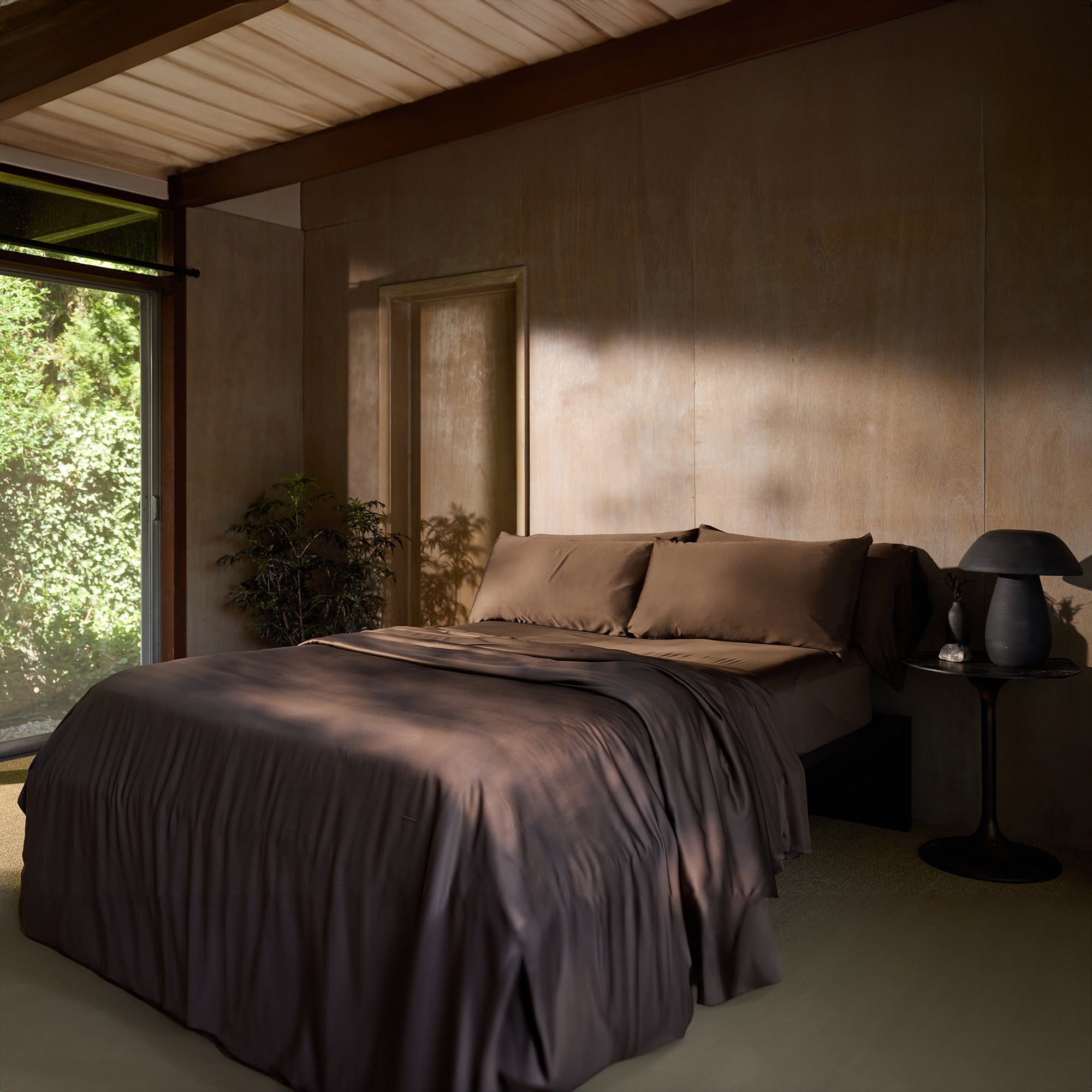 A serene bedroom features a bed adorned with Cozy Earth's Bamboo Sheet Set, flanked by a small black side table with a matching black lamp. Sunlight filters in through a window, casting shadows on a beige wall. A potted plant adds a touch of greenery to the tranquil space. 