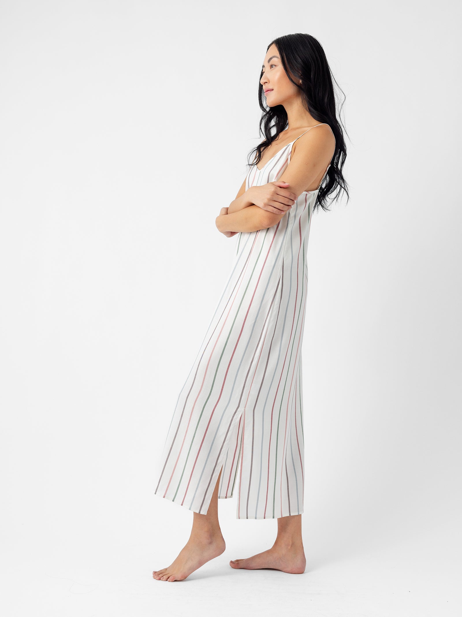 A person with long dark hair is barefoot on a white background, wearing the Cozy Earth Women's Soft Woven Nightgown. They are facing sideways with arms crossed, looking ahead thoughtfully. 