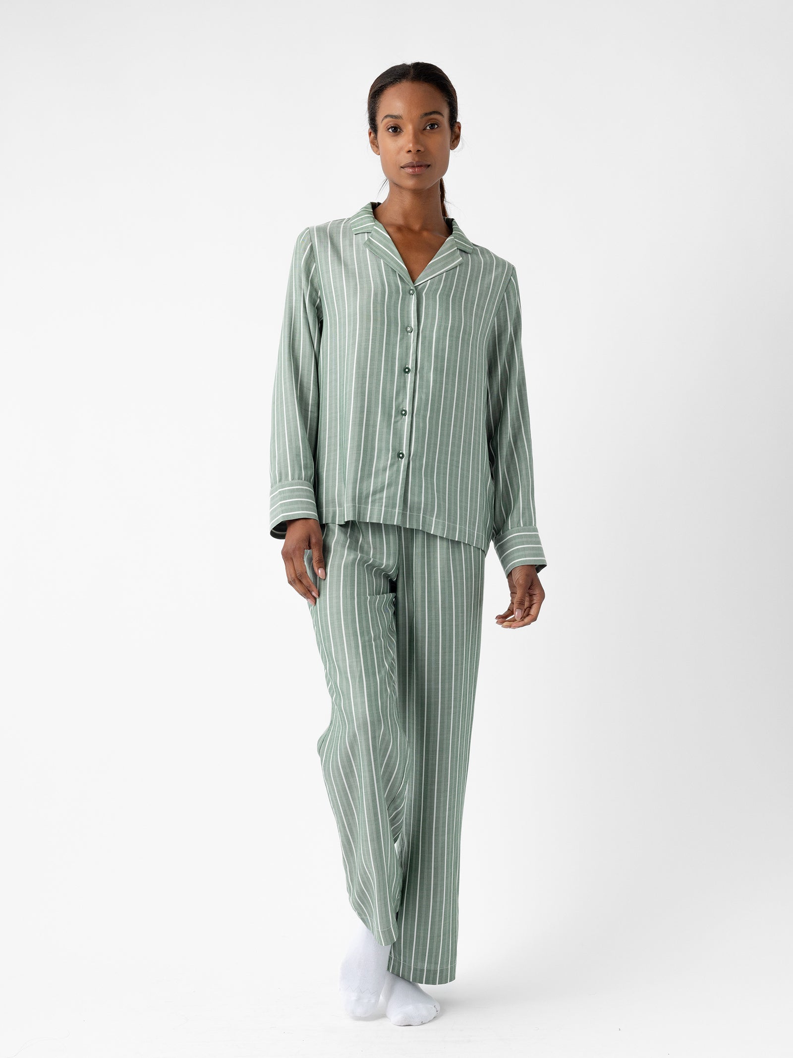 A person is standing against a plain white background, wearing the green and white striped Women's Soft Woven Pajama Pant by Cozy Earth. They have a relaxed pose with their hands by their sides, dressed in a long-sleeve button-up top paired with the matching pants. 