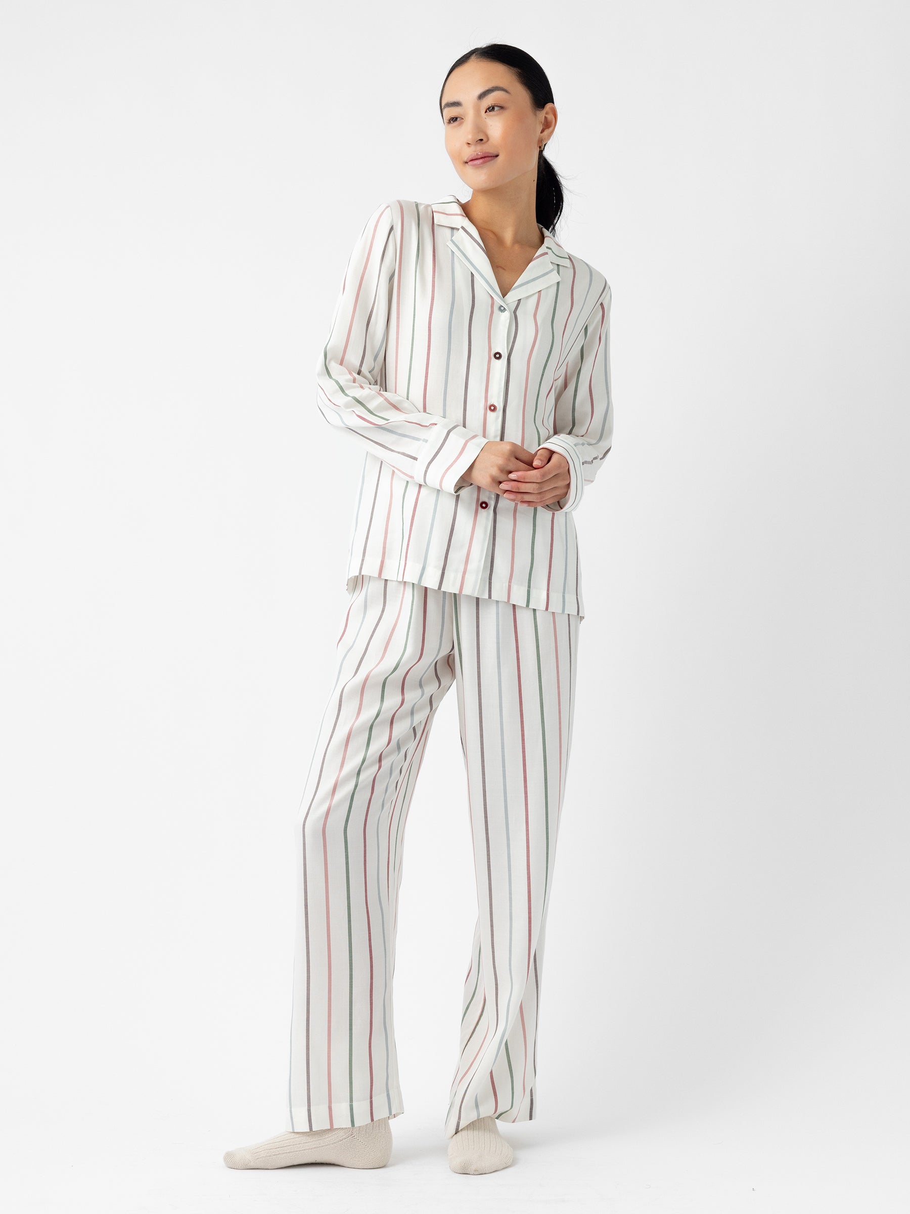 A woman clad in Cozy Earth's Women's Soft Woven Pajama Pant, featuring a design of vertical multicolored stripes on white, stands against a plain backdrop. Her hands rest at her sides as she gazes forward with a neutral expression, pairing the outfit with white socks. |Color:Cottage Stripe