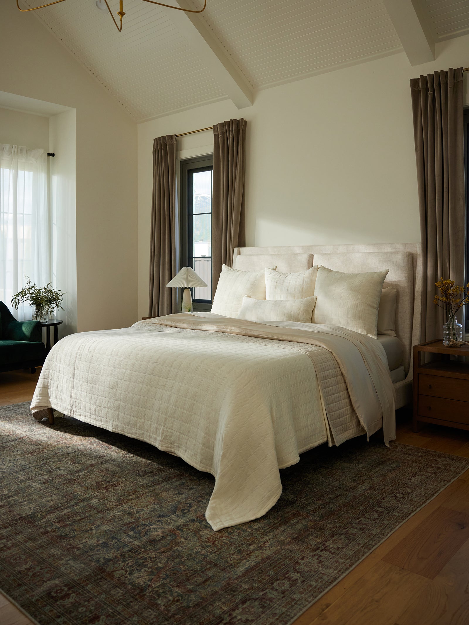 A cozy bedroom with an inviting bed covered in a Waffle Windowpane Blanket by Cozy Earth and pillows. The room features large windows with beige curtains, a wooden nightstand with a lamp and decor, and a patterned rug on a wooden floor. Natural light fills the space, creating a warm ambiance. 