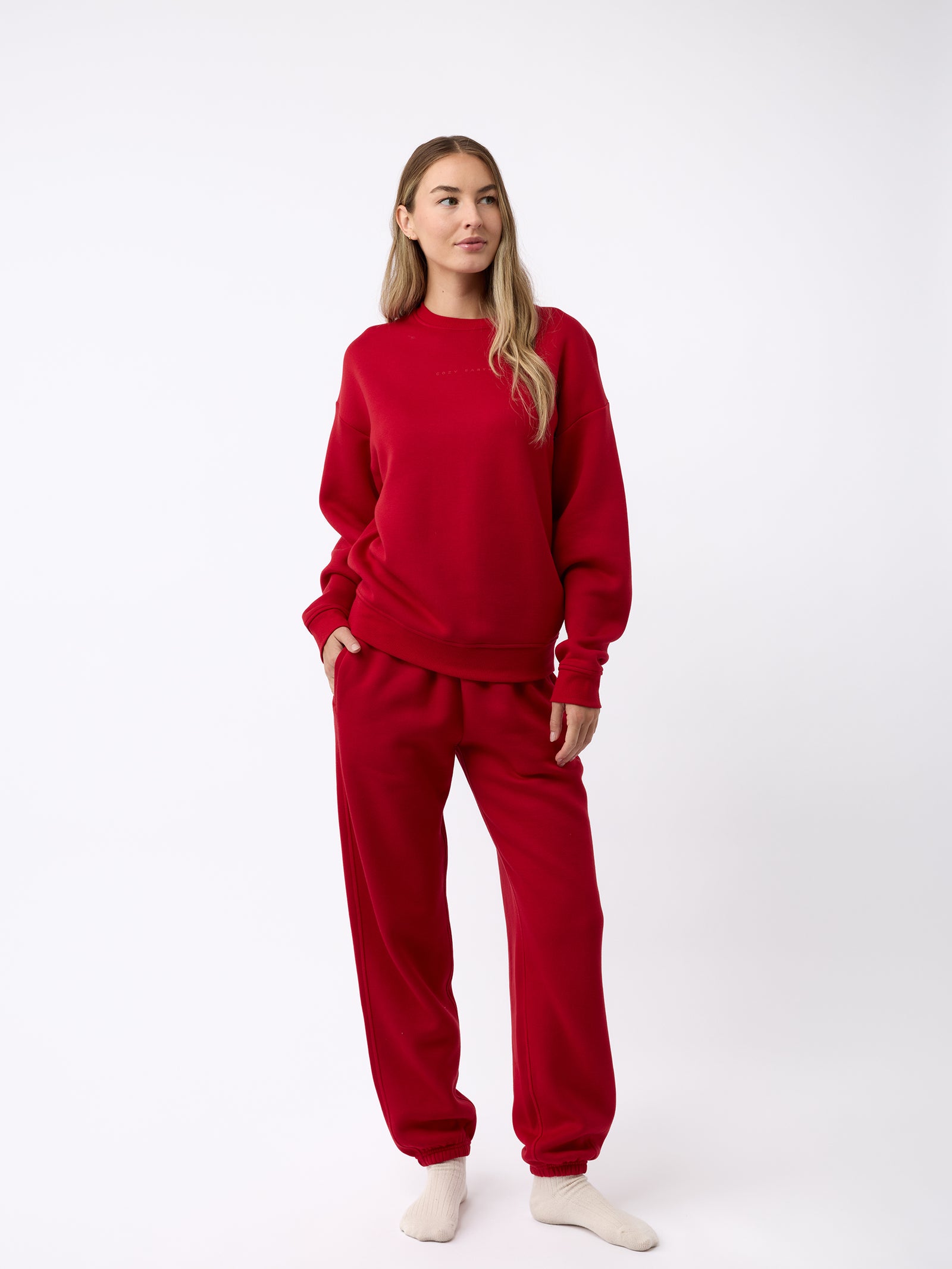 A person wearing Cozy Earth's Women's CityScape Crewneck and matching red sweatpants stands against a plain white background. With long hair, they are looking slightly to the side and have light-colored socks on as well. 