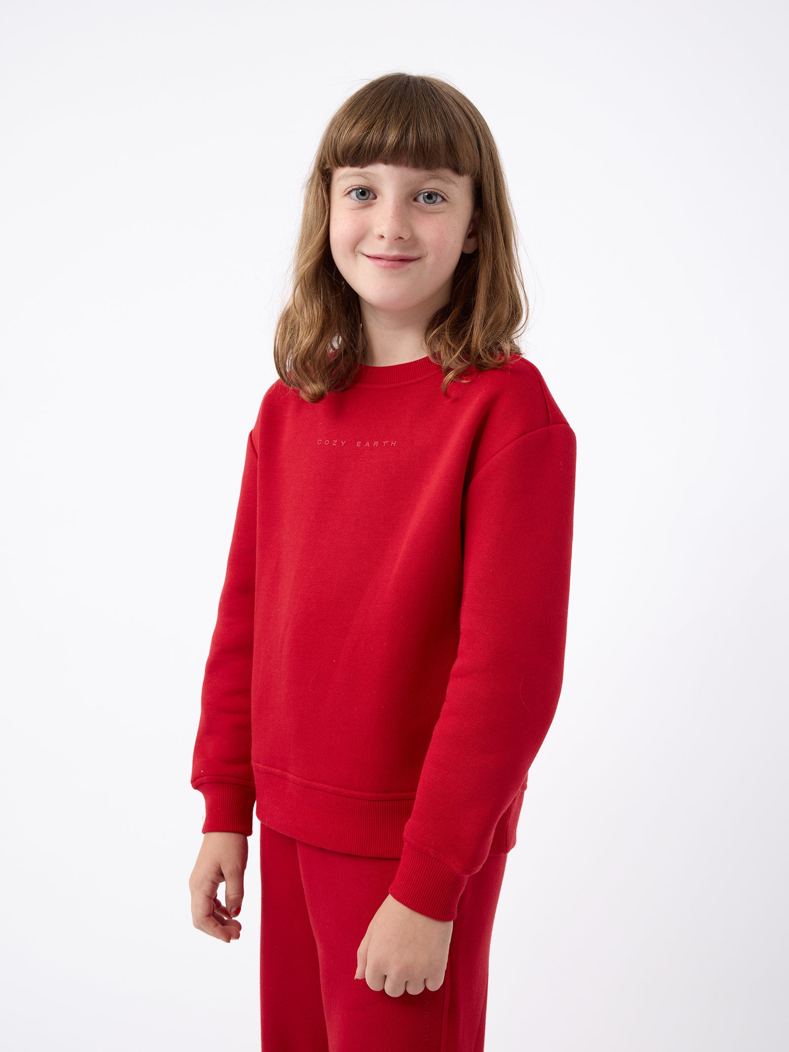 A cheerful young child with brown hair stands against a simple white background, sporting the Kid's CityScape Crewneck and matching pants from Cozy Earth. 