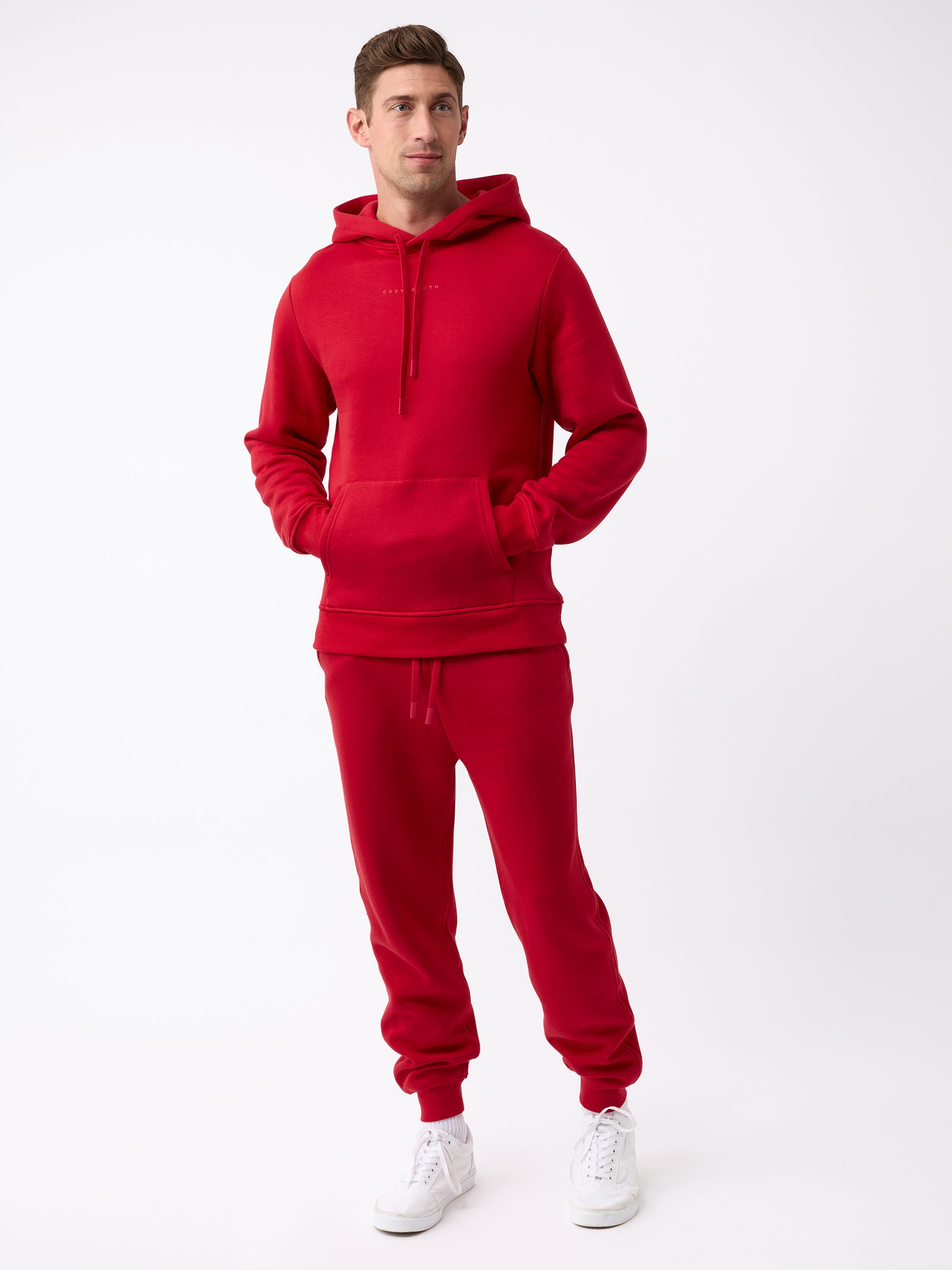 A person in a red Cozy Earth Men's CityScape Hoodie and matching sweatpants stands against a plain white background. They are wearing white sneakers and have their hands in the hoodie pockets, looking slightly to the side. 