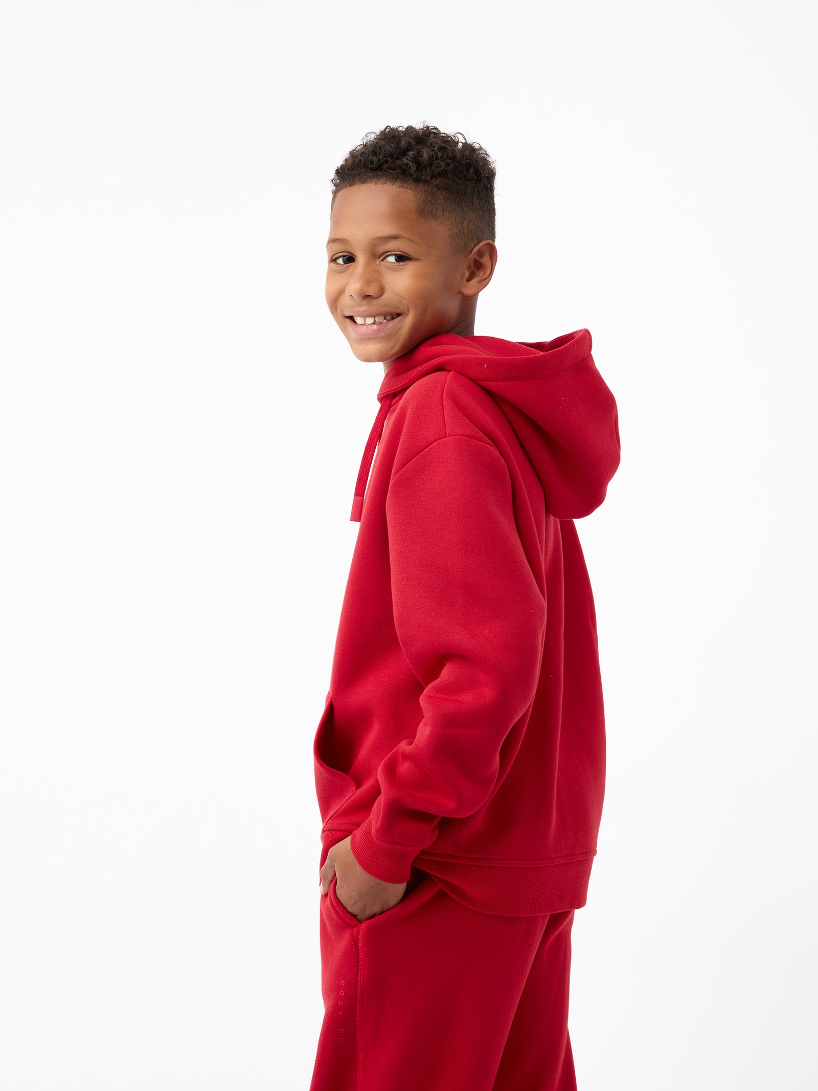 A boy wearing the Kid's CityScape Hoodie from Cozy Earth and matching pants is smiling while standing in a side profile against a white background. 