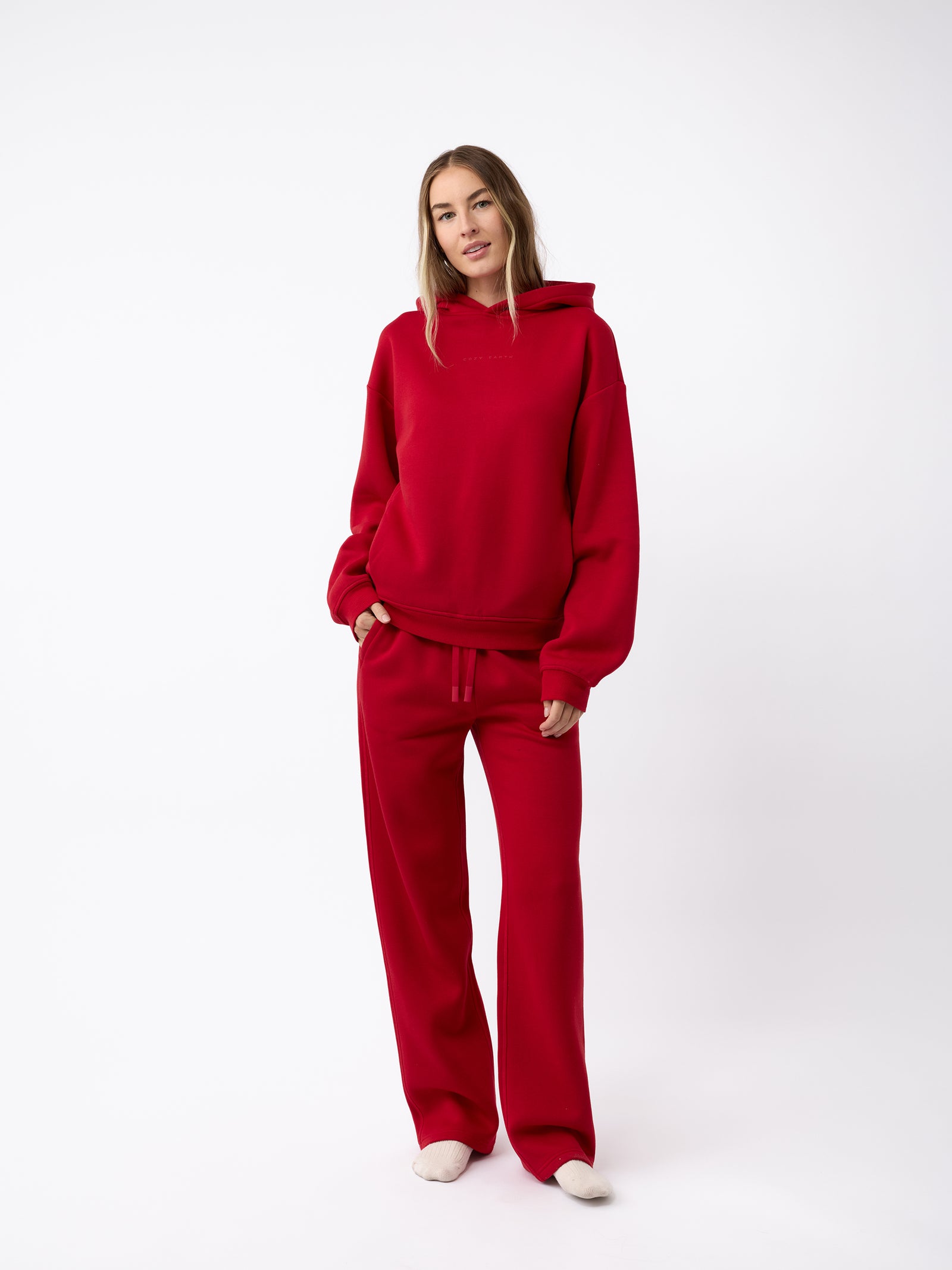 A person stands against a white background wearing a red hoodie with matching Cozy Earth's Women's CityScape Wide Leg Pant in red. They have long hair and are looking at the camera with a relaxed pose while wearing white shoes. 