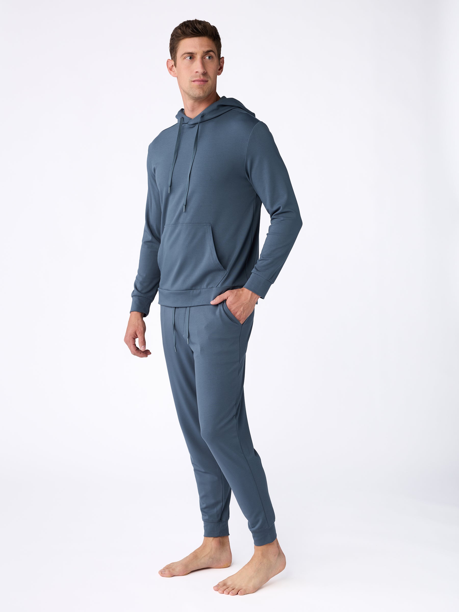 A man stands barefoot against a white background, dressed in Cozy Earth's Men's Ultra-Soft Bamboo Hoodie and matching jogger pants. His hands rest in his pockets as he gazes slightly to the side. |Color:Dawn