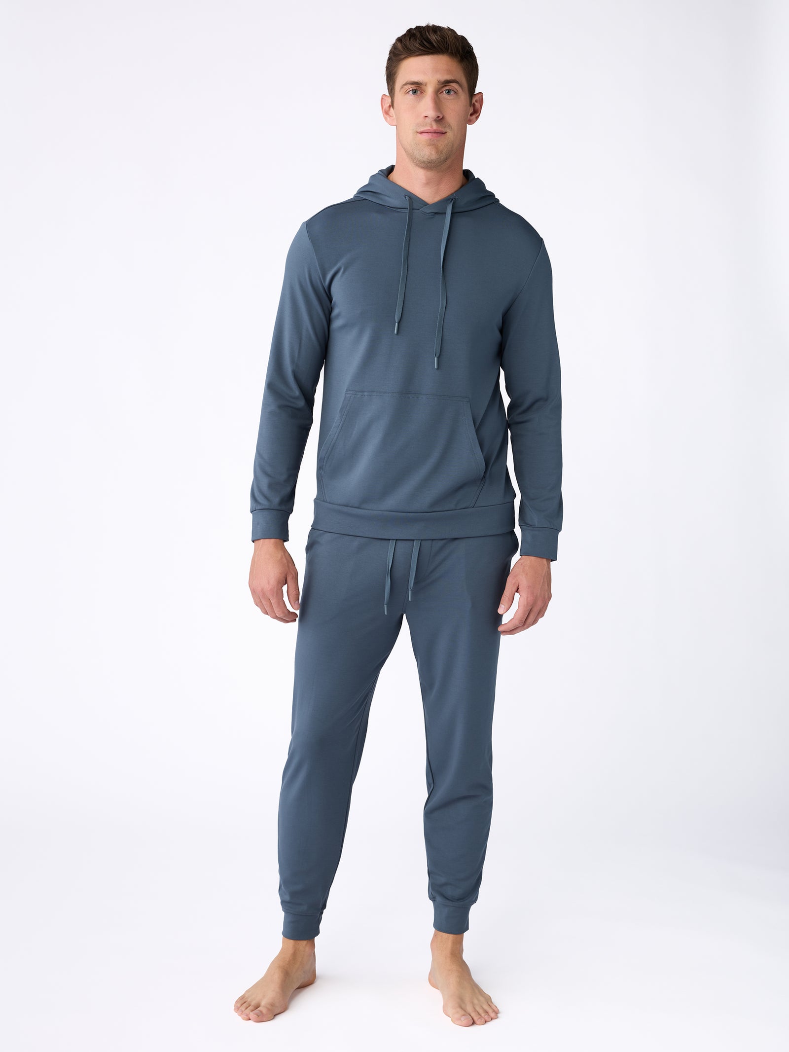 A man with short, dark hair and a neutral expression stands barefoot on a white background, dressed in Cozy Earth's Men's Ultra-Soft Bamboo Pullover Crew in teal. The pullover features a front pocket and drawstrings, while the matching jogger pants are fitted and comfortable. 