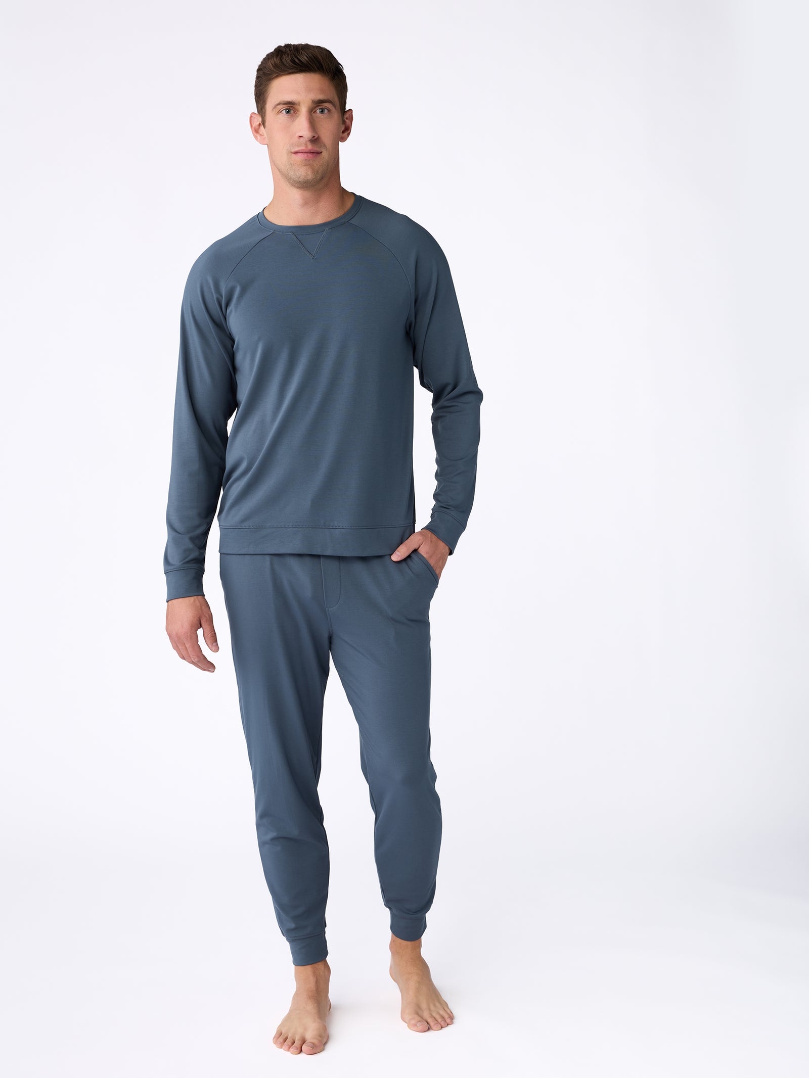 A person stands barefoot against a plain white background, sporting a matching light blue athletic ensemble featuring the Men's Ultra-Soft Bamboo Pullover Crew by Cozy Earth and jogger-style pants. 