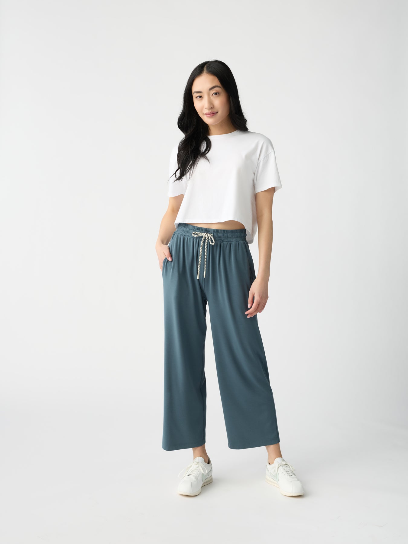 A person with long black hair stands against a plain background. They wear a white crop top, Cozy Earth's Women's Studio Cropped Wide Leg Pant in blue with a drawstring, and white sneakers. Their posture is relaxed with hands in pockets. |Color:Dawn