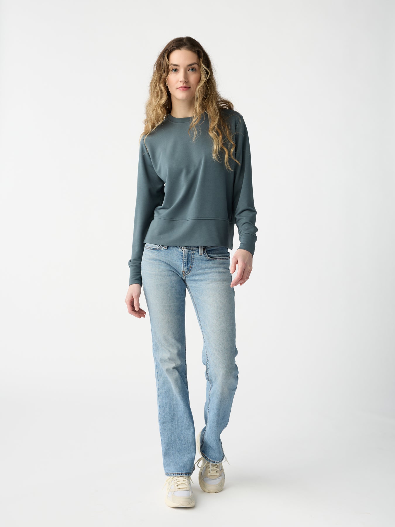 A person with long wavy hair is wearing a teal Women's StudioLite Crewneck from Cozy Earth, light blue jeans, and white sneakers. They stand against a plain white background, looking directly at the camera. 