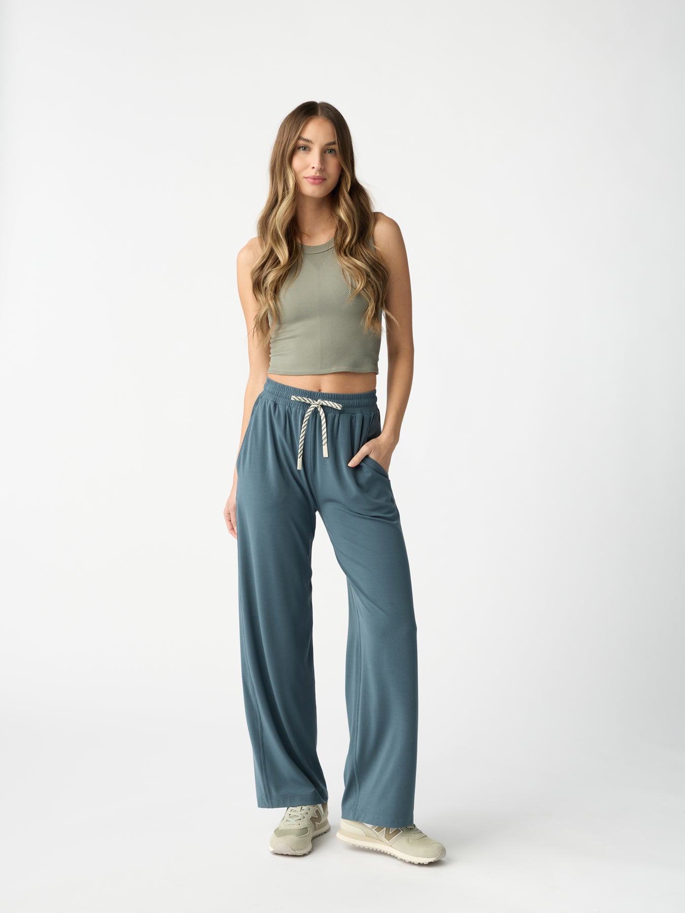 A woman with long hair wears a green sleeveless top and Cozy Earth's Women's Studio Wide Leg Pant, featuring a drawstring waist. She stands casually with her hands in her pockets against a plain, light background. |Color:Dawn