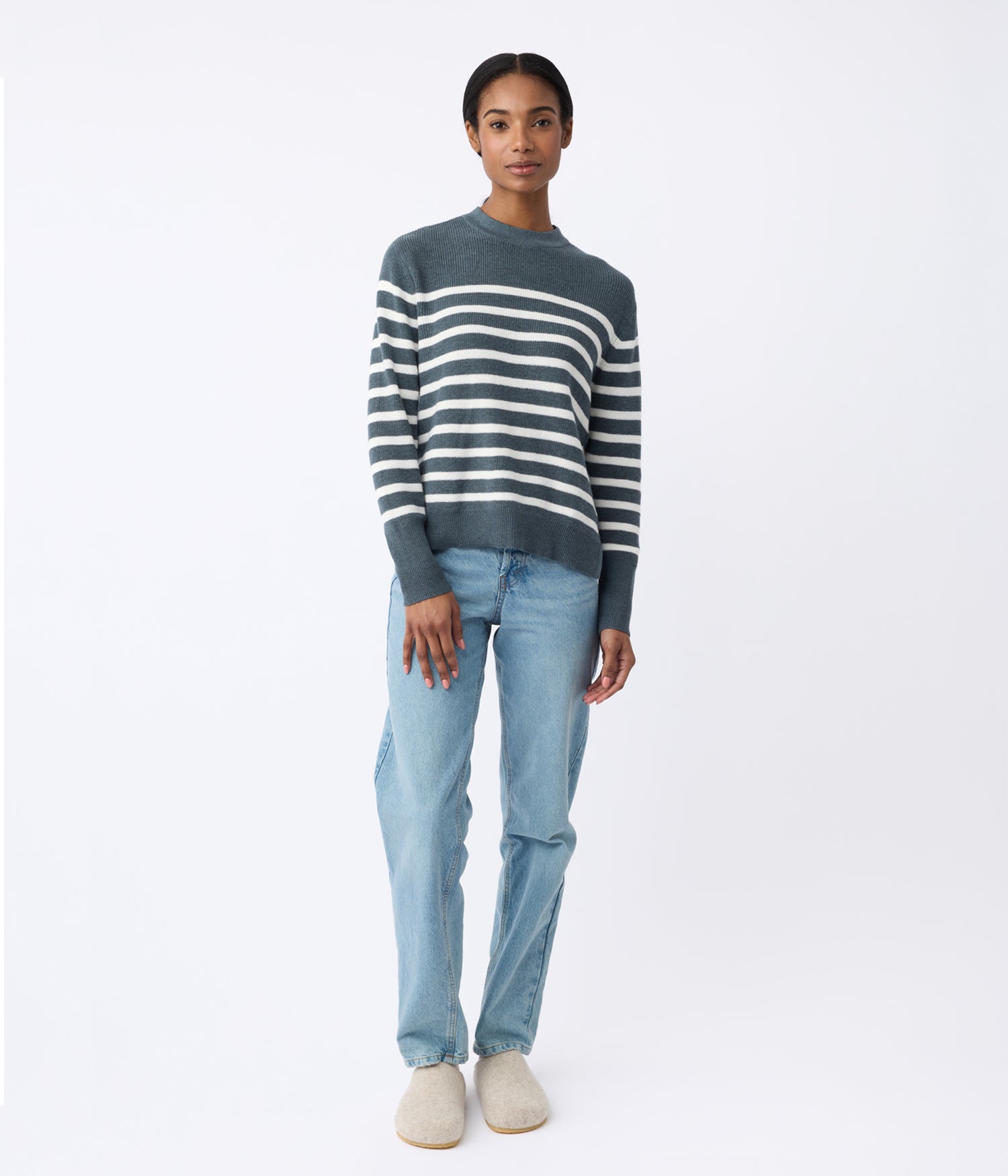 A person stands against a white background wearing Cozy Earth's Women's Rowan Sweater in gray and white stripes, paired with light blue jeans and beige shoes. |Color:Dawn/Bone Stripe