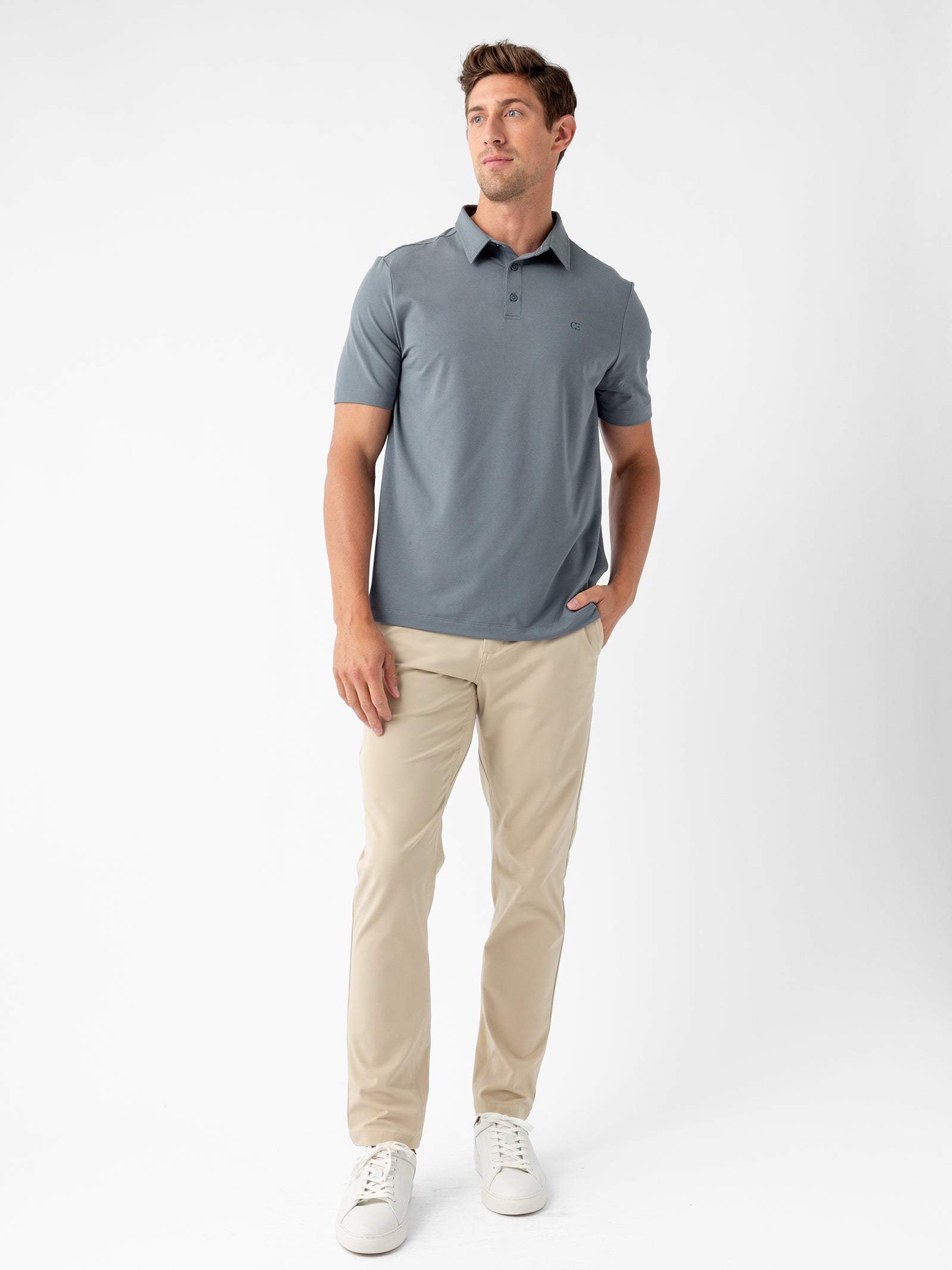 A man stands against a plain white background, wearing Cozy Earth's Men's Everyday Polo in slate blue with beige pants. His right hand rests in his pant pocket, and he gazes slightly off to the side. He complements his outfit with white sneakers and maintains a neutral expression. 