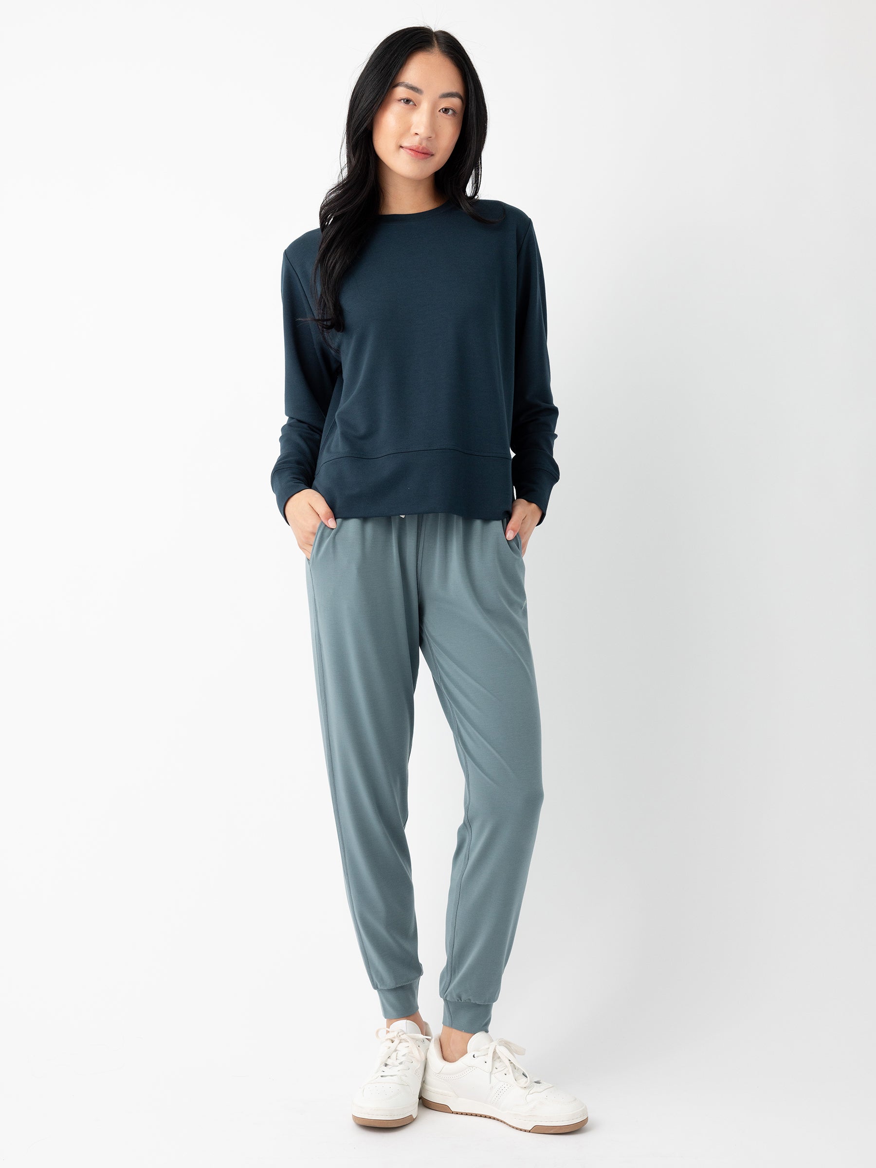 A person with long black hair, wearing a dark blue long-sleeve shirt and light blue Women's Studio Jogger pants by Cozy Earth, is standing against a plain white background. Their hands are tucked into the pockets of the pants, and they are wearing white sneakers. |Color:Deep Blue