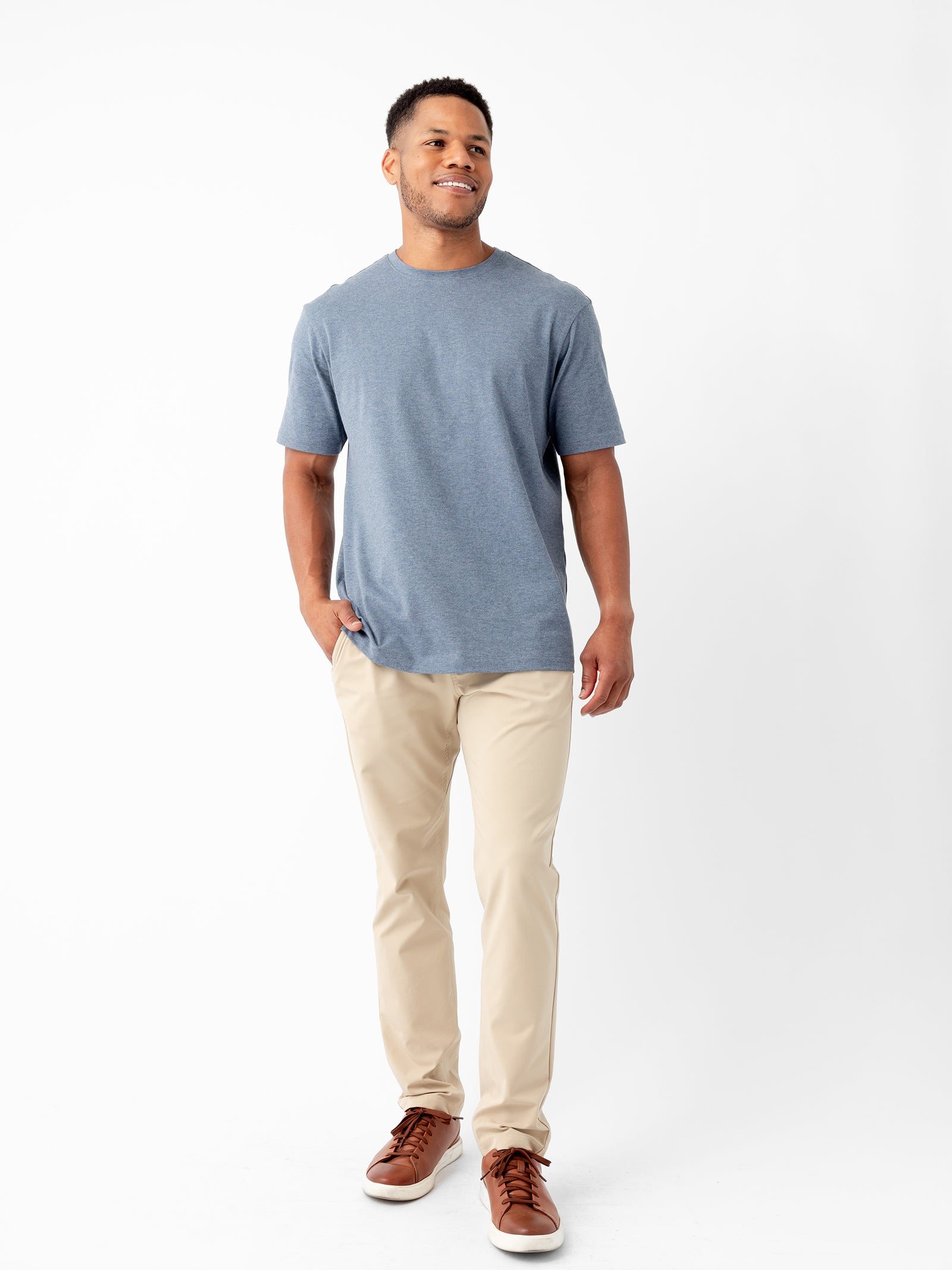 A man stands against a plain white background. He is smiling and wearing the Cozy Earth Men's All Day Tee in light blue, beige pants, and brown sneakers. His left hand is in his pocket while his right arm hangs by his side. 