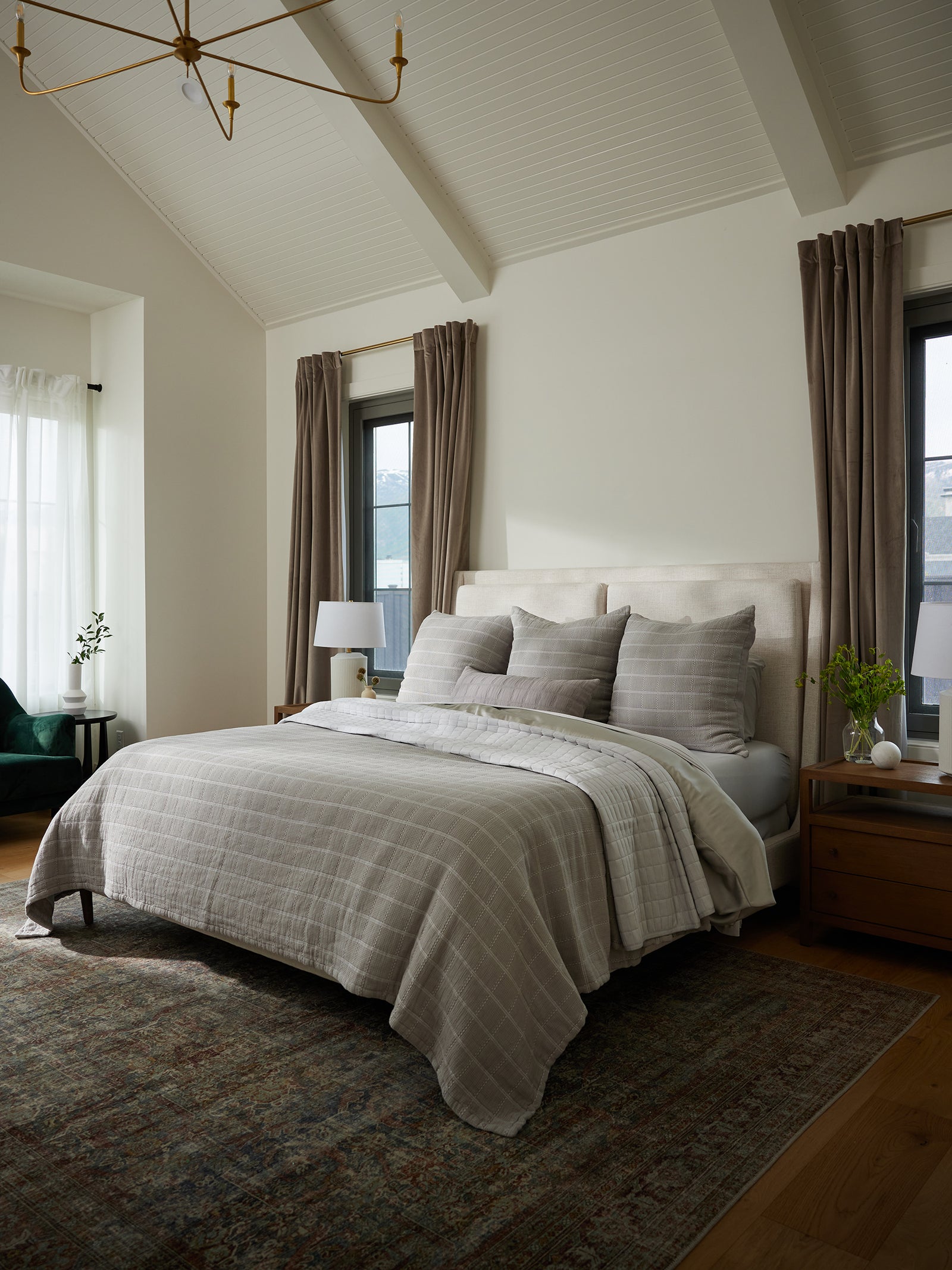 A cozy bedroom with a large bed draped in a Cozy Earth Waffle Windowpane Blanket, featuring neutral-toned bedding and pillows. Two windows with beige curtains frame the bed, and on each side, there is a wooden nightstand adorned with a lamp and a small potted plant. A patterned rug blankets the hardwood floor. 