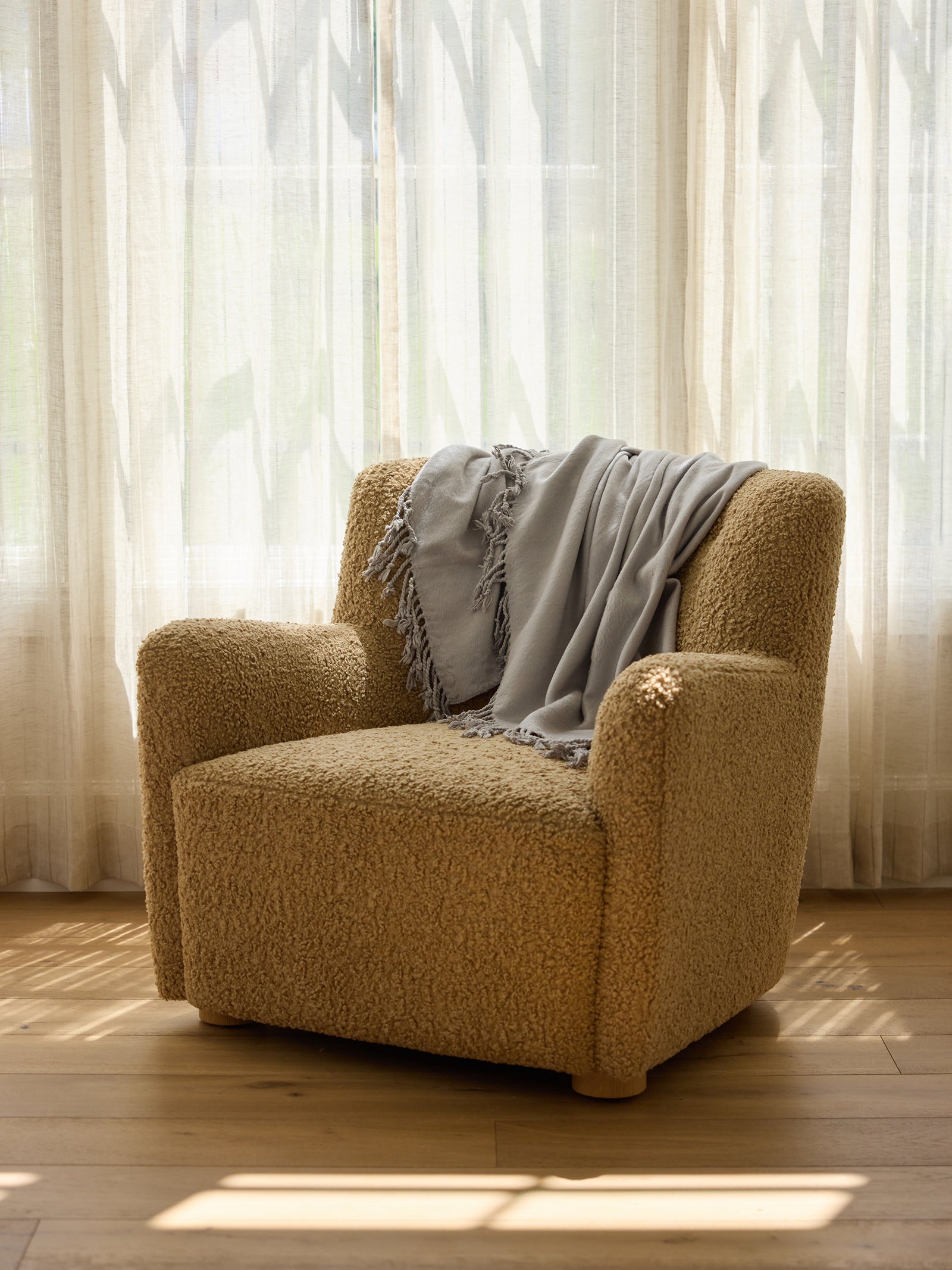 A cozy beige armchair with textured fabric is placed in front of a sheer, sunlit curtain. A light gray Bamboo Tassel Throw by Cozy Earth is draped casually over the backrest, and sunlight casts soft shadows on the wooden floor. 