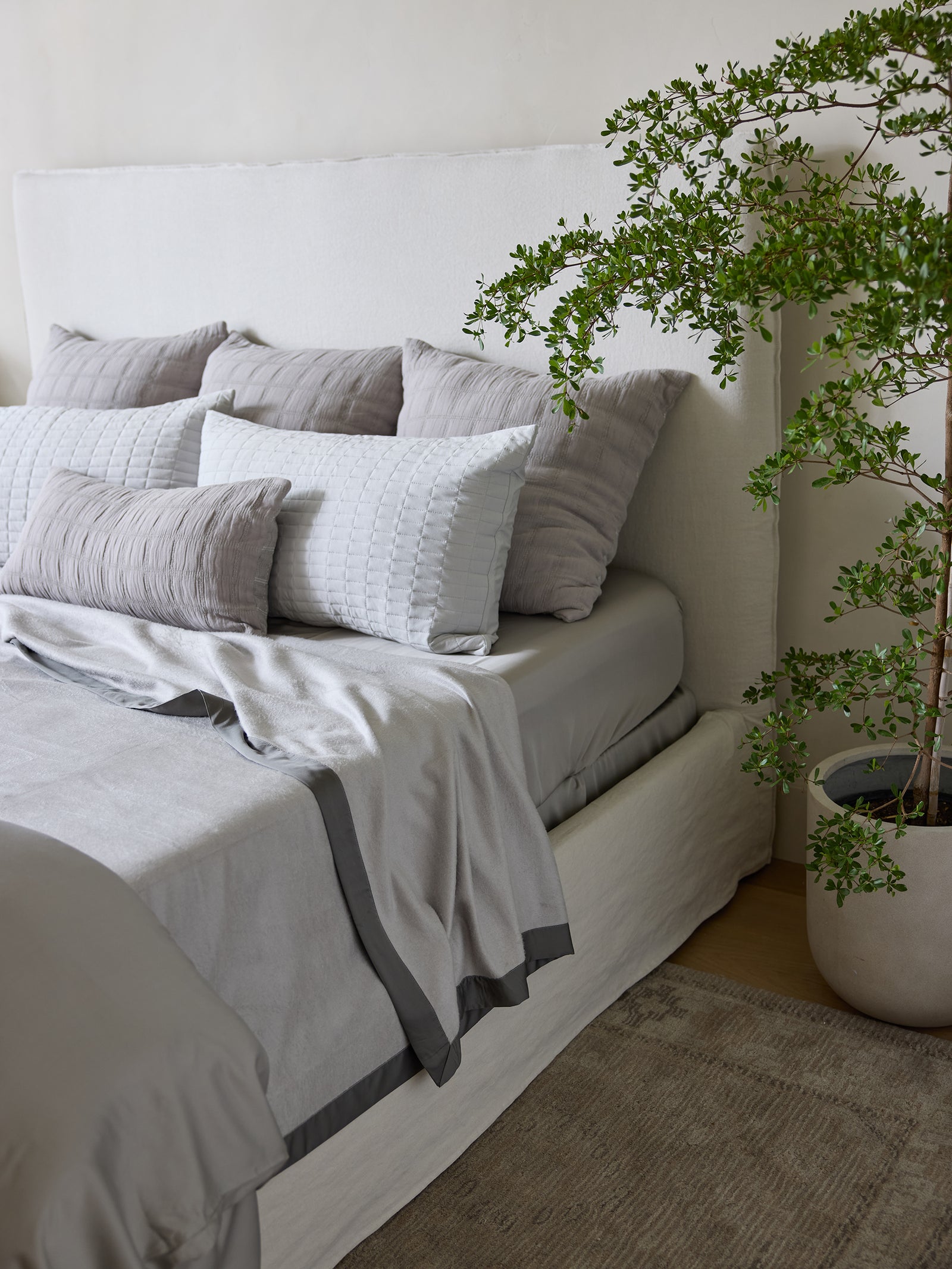 A neatly made bed with a white upholstered headboard, gray pillows, and a Cozy Earth Bamboo Blanket in light gray partially folded down. A potted green plant is placed next to the bed, adding a touch of nature to the serene bedroom setting. The floor is adorned with a beige rug. 