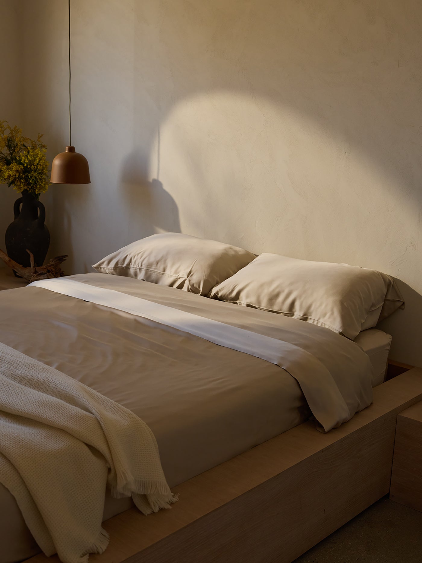 A minimalist bedroom features a neatly made bed with a beige Bamboo Duvet Cover by Cozy Earth. A brown pendant light hangs above a wooden nightstand, beside which stands a dark vase with yellow flowers. Soft sunlight casts shadows on the cream-colored wall. |Color:Driftwood