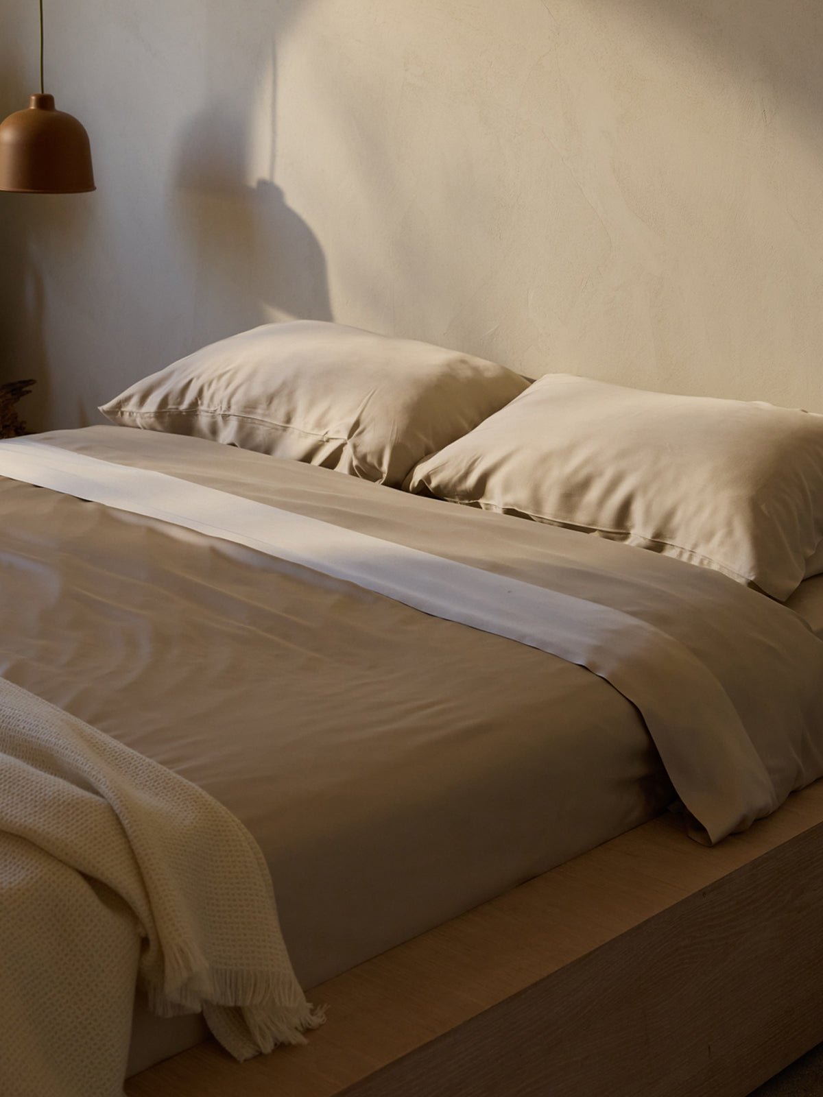 A neatly made bed with the Cozy Earth Bamboo Bedding Deluxe Bundle, featuring beige sheets and matching pillows. Soft lighting from a nearby lamp casts a warm glow, creating a cozy atmosphere. A textured white blanket is draped over one corner of the bed. 