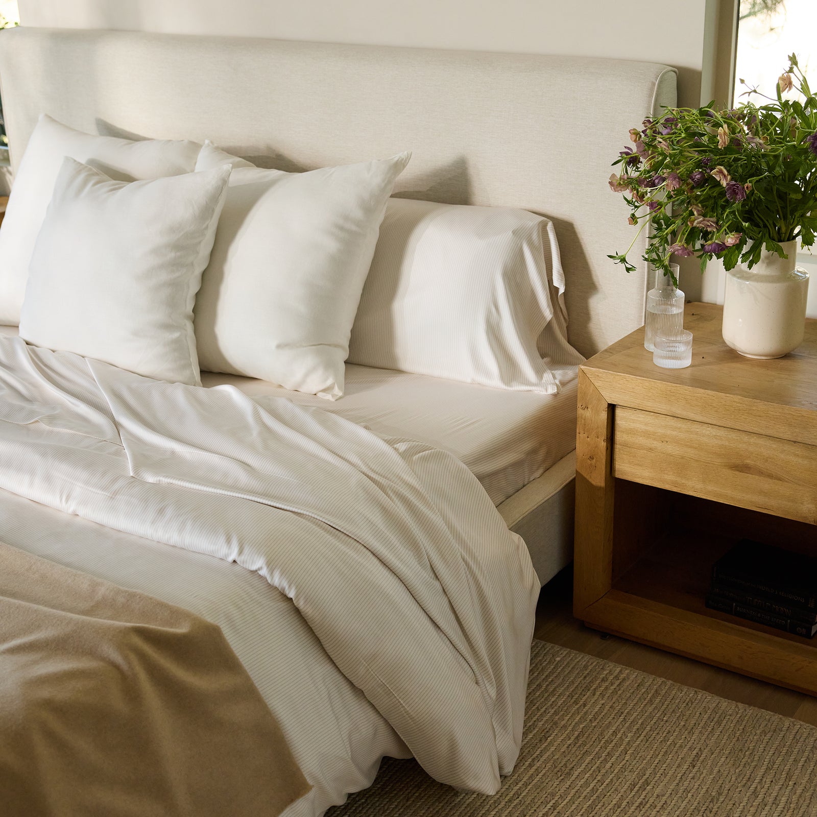 A cozy bedroom scene featuring a bed adorned with Cozy Earth's Bamboo Duvet Cover, complemented by white pillows and sheets, a beige blanket, and a light beige headboard. A wooden nightstand holds a potted plant and a glass of water, with books neatly stacked below. A window with greenery is in the background. 