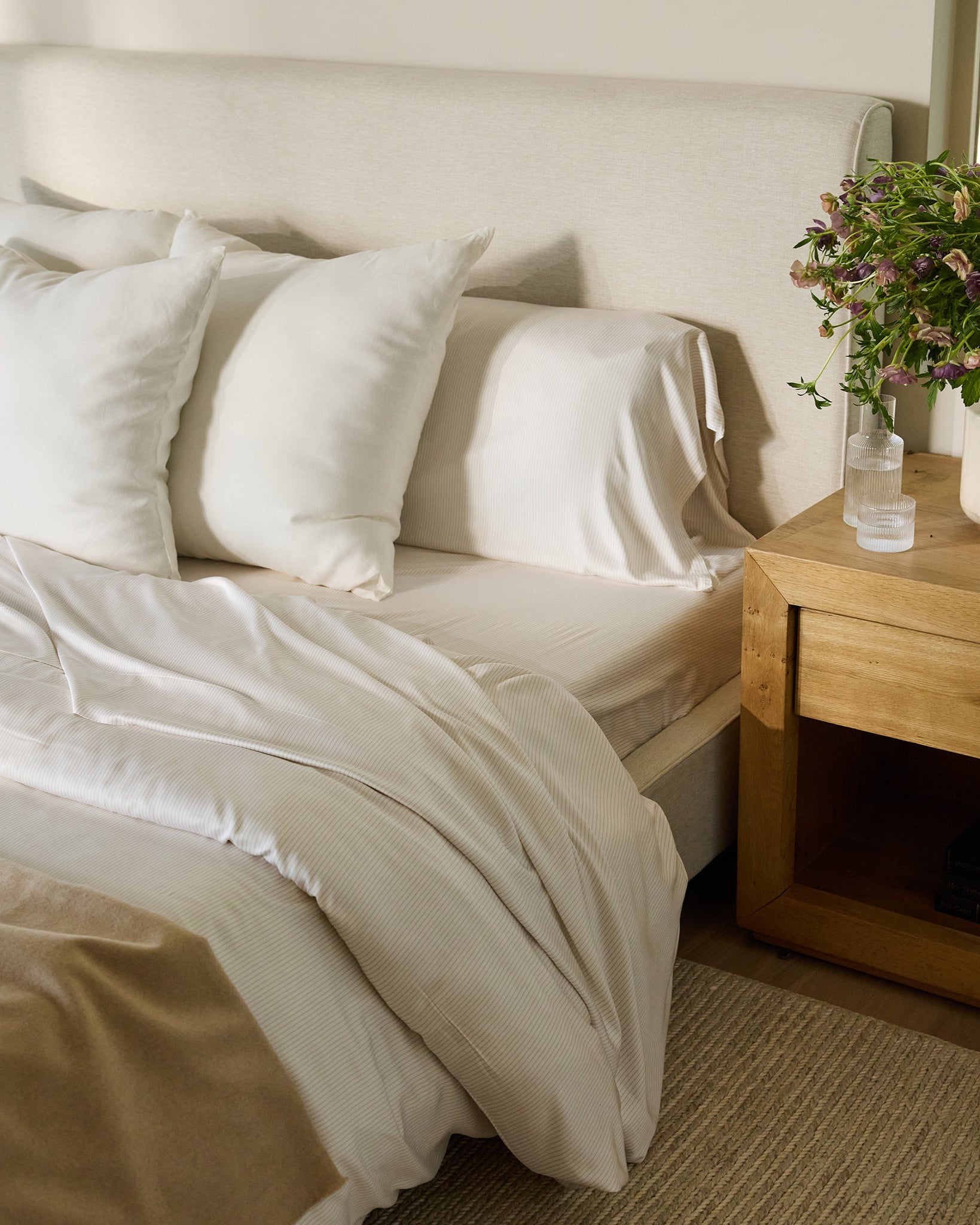 A cozy bedroom scene featuring a bed adorned with Cozy Earth's Bamboo Duvet Cover, complemented by white pillows and sheets, a beige blanket, and a light beige headboard. A wooden nightstand holds a potted plant and a glass of water, with books neatly stacked below. A window with greenery is in the background. 