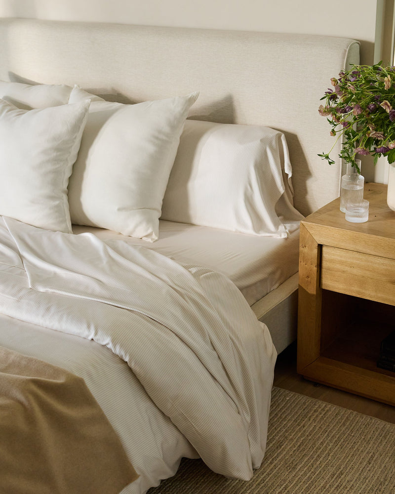 A cozy bedroom scene featuring a bed adorned with Cozy Earth's Bamboo Duvet Cover, complemented by white pillows and sheets, a beige blanket, and a light beige headboard. A wooden nightstand holds a potted plant and a glass of water, with books neatly stacked below. A window with greenery is in the background.