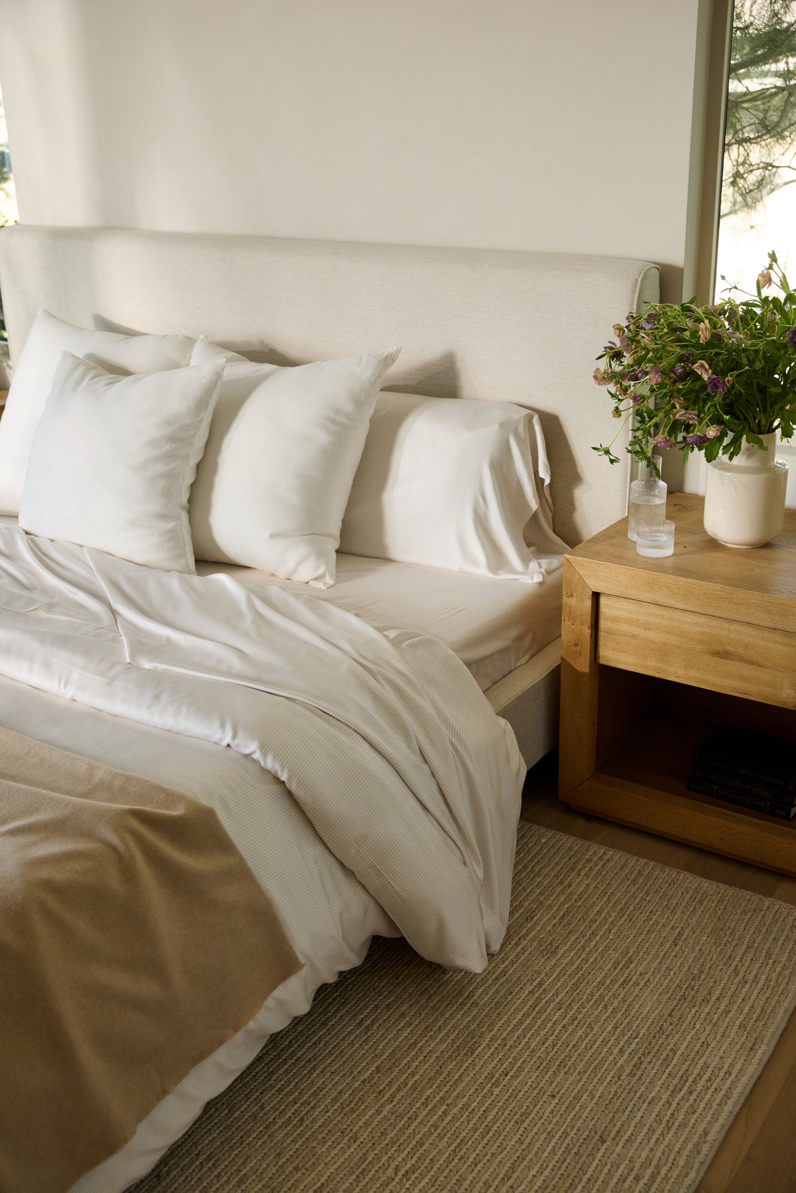 A cozy bedroom with a neatly made bed showcasing Cozy Earth's Bamboo Fitted Sheet along with white pillows. A wooden nightstand holds a potted plant and a glass of water. Natural light filters through an unseen window, casting a soft glow across the room. 