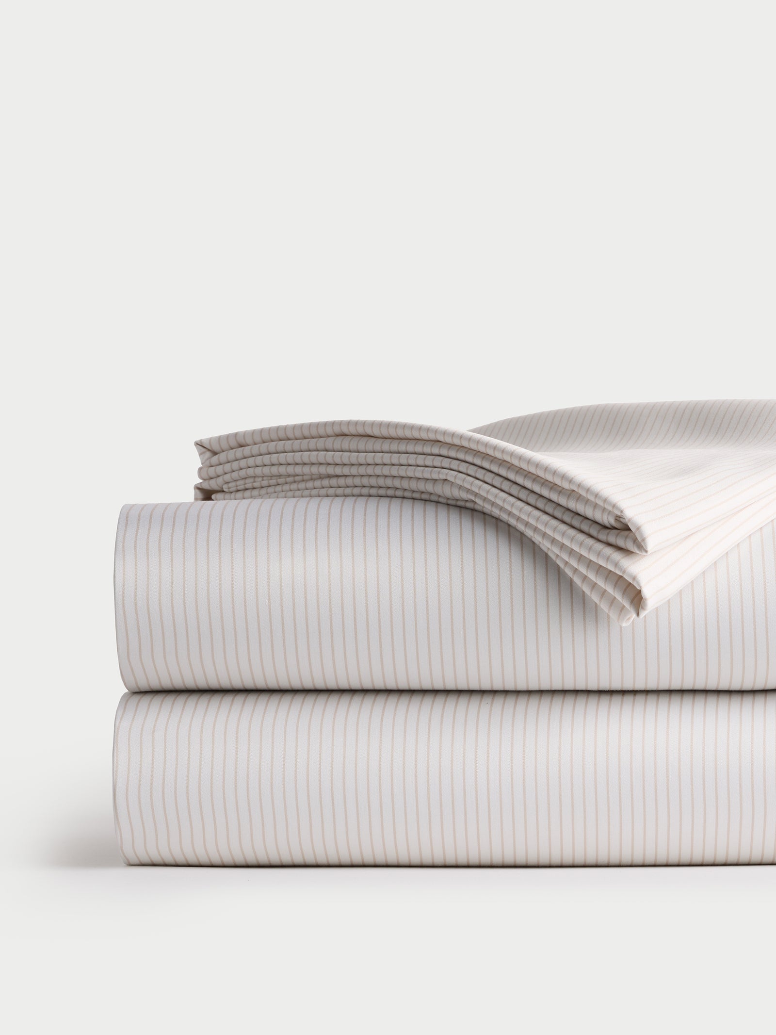 A neatly folded stack of Cozy Earth's Bamboo Sheet Set in neutral tones, featuring striped bed sheets and pillowcases. The fabric is adorned with thin vertical stripes that create a subtle pattern, while the plain, light background accentuates the clean and organized presentation. 