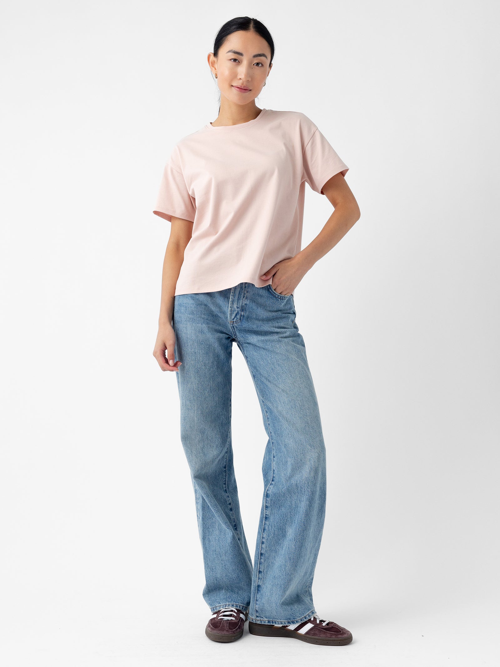 A woman stands confidently against a plain white background. She is wearing Cozy Earth's Women’s All Day Tee in light pink, paired with blue wide-leg jeans and maroon sneakers featuring white stripes. Her right hand rests on her hip while her left hand hangs loosely by her side. 