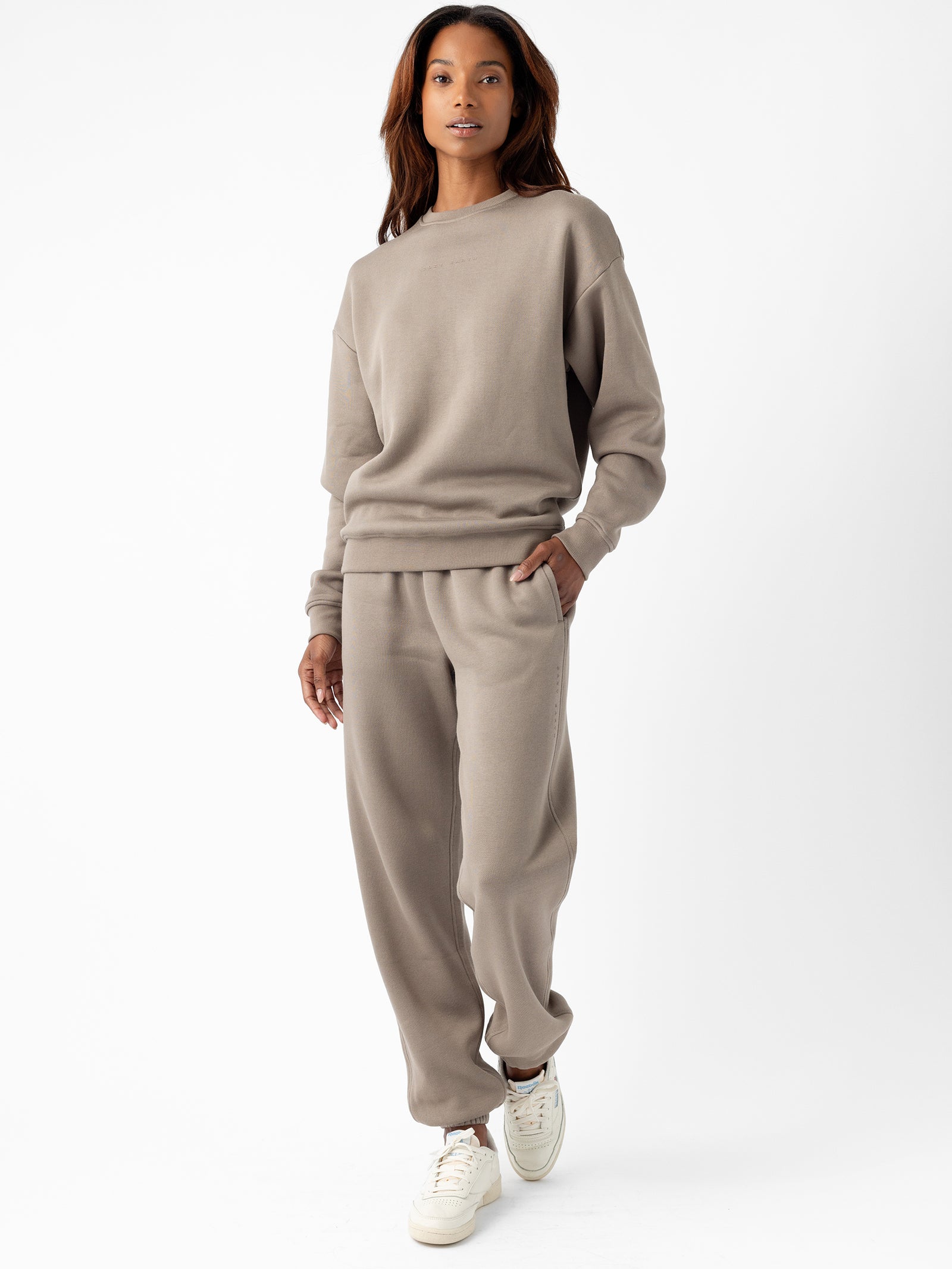 A person with long, dark hair wearing a matching beige Women's CityScape Crewneck and sweatpants from Cozy Earth stands with one hand in their pocket. They are also wearing white sneakers and are posed against a plain white background. 