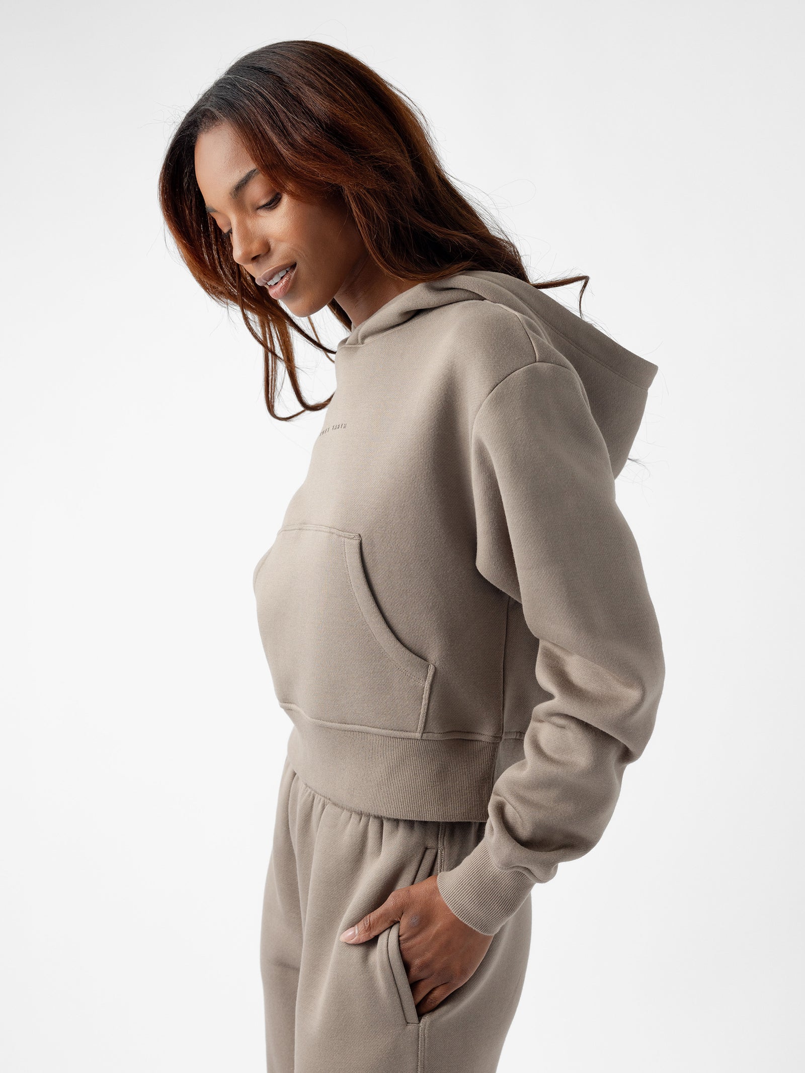 A person with long hair is wearing Cozy Earth's Women's CityScape Cropped Hoodie in a taupe color along with matching sweatpants. They are looking down and slightly to the side, with one hand in their pocket against a white background. 