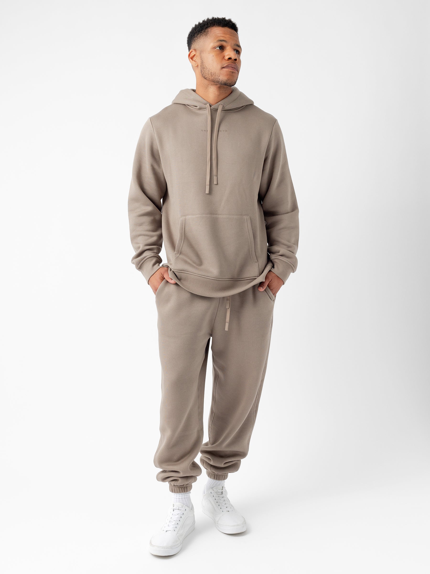 A man stands against a plain white background wearing the taupe-colored Men's CityScape Hoodie and matching jogger pants by Cozy Earth, paired with white high-top sneakers. He has his hands in the hoodie pocket and is looking off to the side. |Color:Ebony