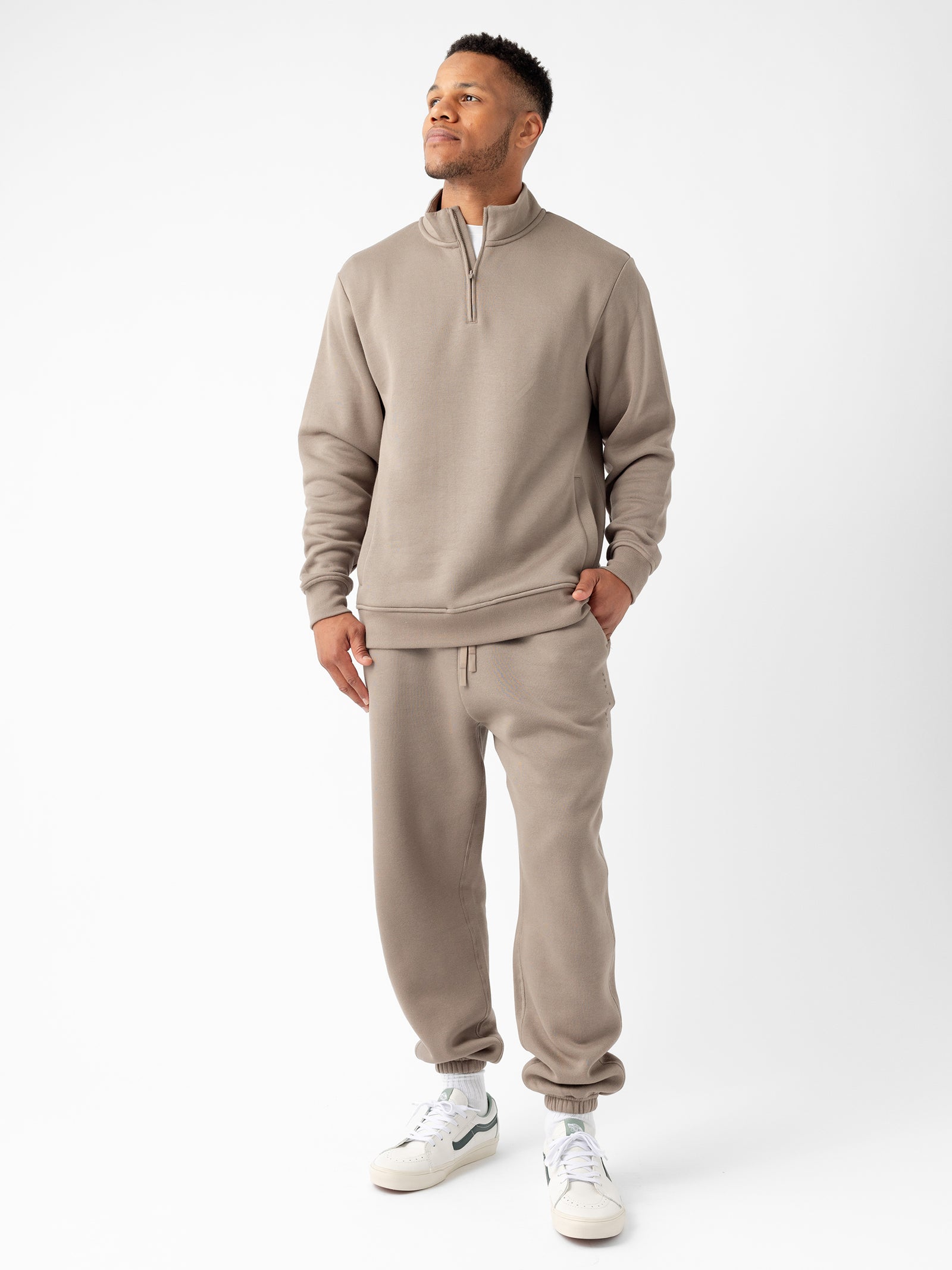 A person stands against a white background, wearing Cozy Earth's Men's CityScape Quarter Zip in taupe-colored fabric and matching sweatpants with pockets. They have their left hand in their pocket and are looking slightly upward, smiling. The outfit is paired with white sneakers. 