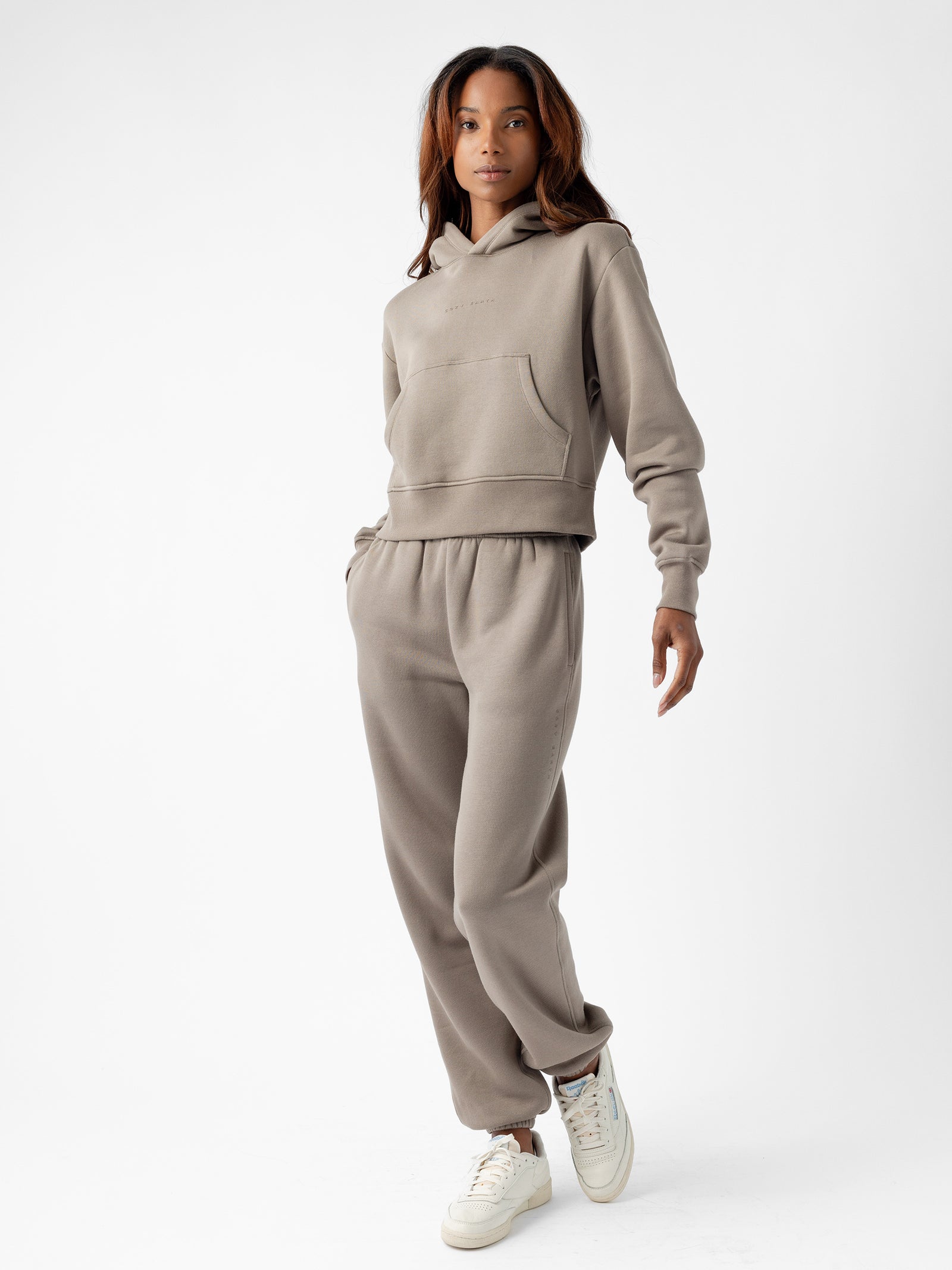 A person wearing a Cozy Earth taupe-colored hoodie and Women's CityScape Sweatpant stands against a white background. They are also wearing white sneakers and have a relaxed pose, with one hand in a pocket and the other arm slightly raised. The person has long hair and a calm expression. 