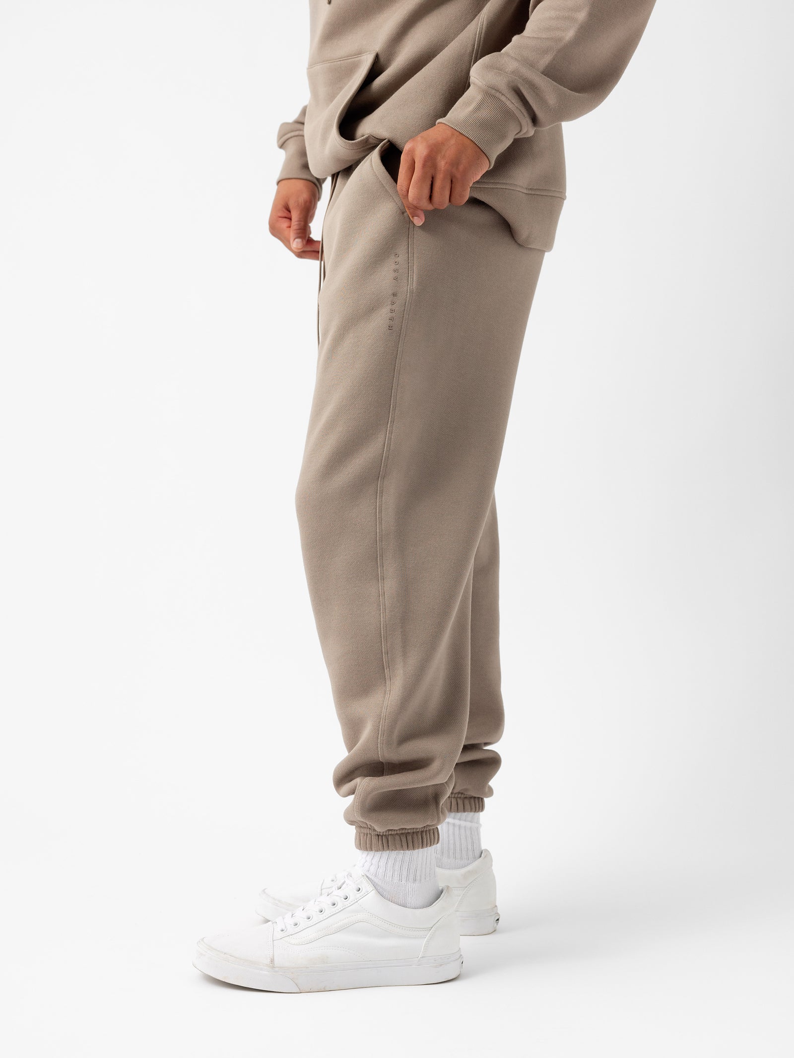 A person is wearing a Men's CityScape Sweatpant by Cozy Earth, along with a matching beige sweatshirt, paired with white sneakers and white socks. The background is plain white, emphasizing the casual and comfortable outfit. The person's face is not visible. 