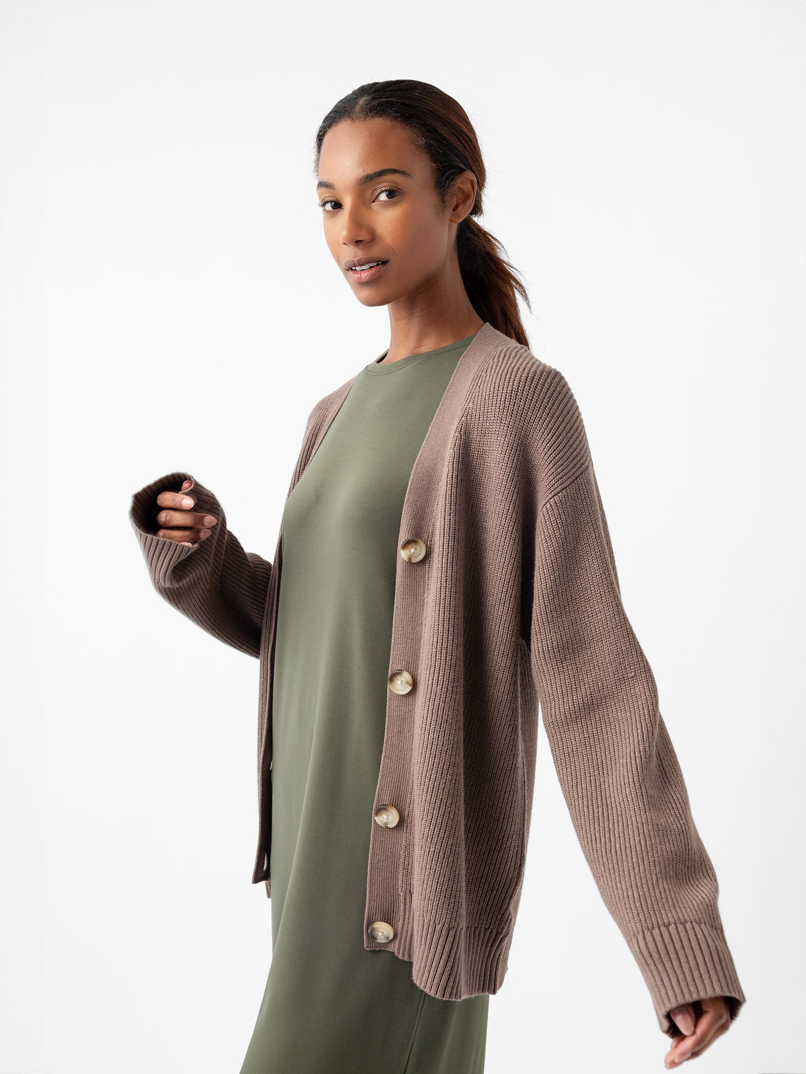 Against a plain white background, an individual is wearing a green dress complemented by the Oversized Classic Cardigan from Cozy Earth, featuring prominent buttons. Their long hair is pulled back as they gaze slightly to the side with a relaxed expression. 