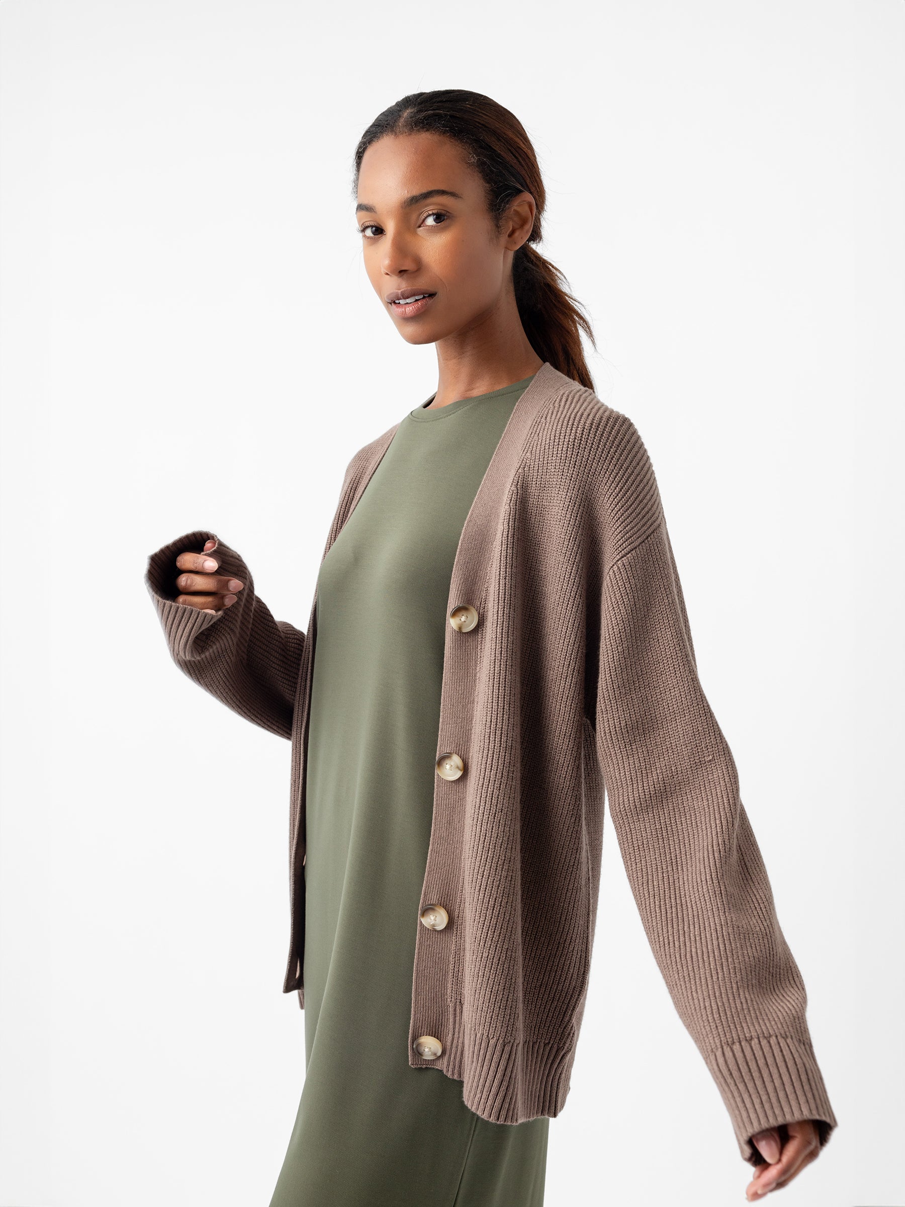 Against a plain white background, an individual is wearing a green dress complemented by the Oversized Classic Cardigan from Cozy Earth, featuring prominent buttons. Their long hair is pulled back as they gaze slightly to the side with a relaxed expression. |Color:Ebony