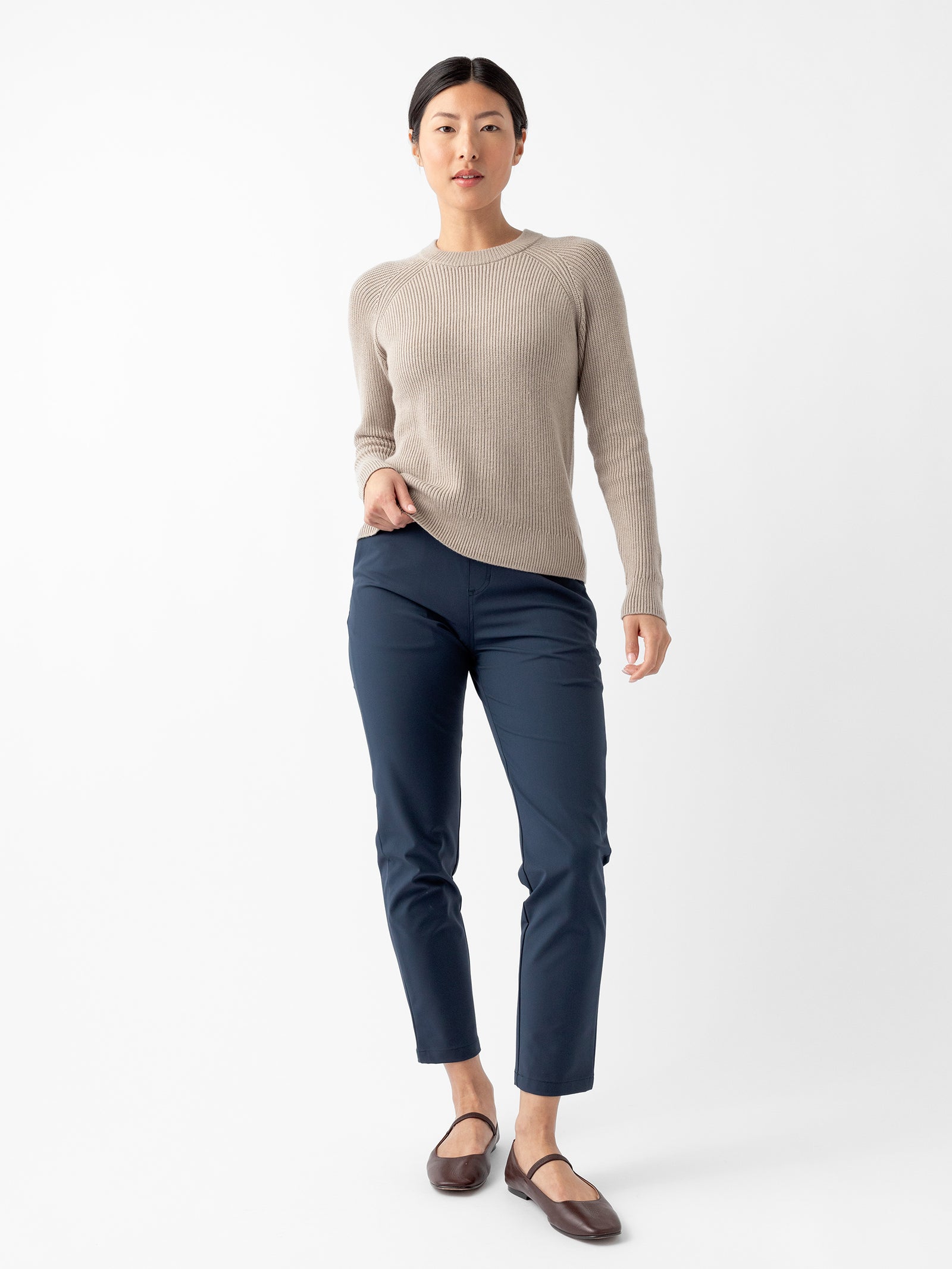 A person with dark hair is standing against a plain white background. They are wearing a beige long-sleeve sweater, Cozy Earth's Women's Always Cropped Pant in navy blue, and brown flats. Their hands are relaxed by their sides and they have a neutral expression. 