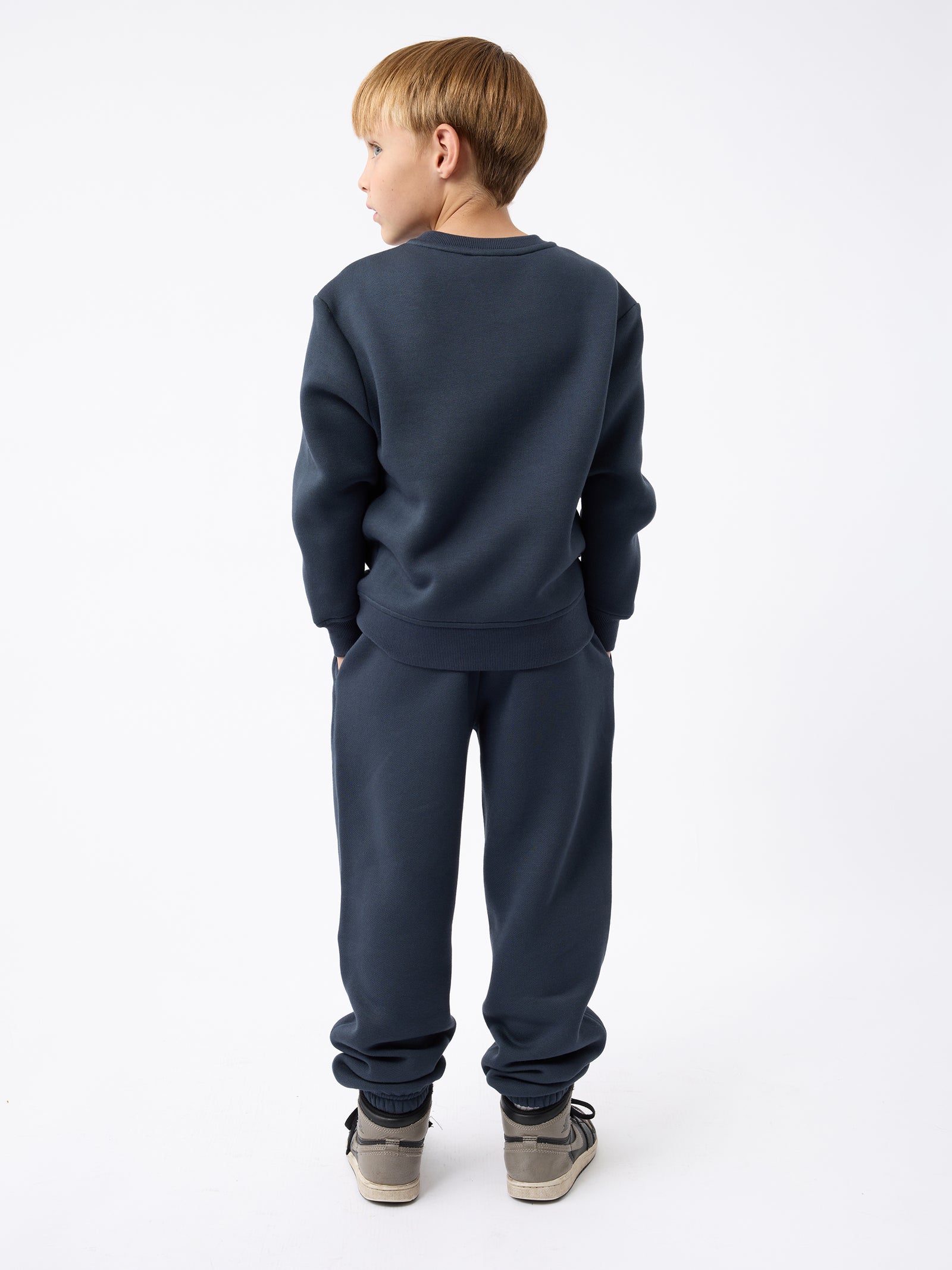 A young boy with blonde hair is standing with his back to the camera, wearing a Kid's CityScape Crewneck in dark blue from Cozy Earth and gray sneakers. His hands are in his pockets, and he is looking slightly to the side against a plain white background. 